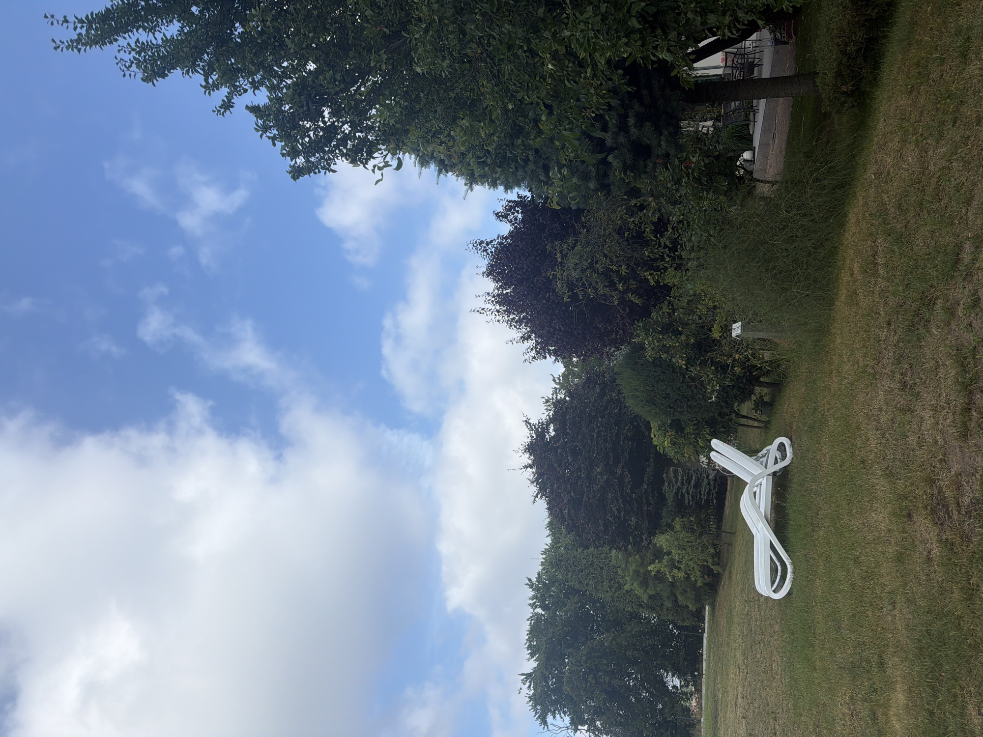 A white plastic lounge chair is situated on a grassy area. Behind the chair, a variety of trees and bushes form a dense line. Beyond the foliage, a patio with chairs and tables is visible, and further in the background, a building can be seen. The sky above is blue with scattered white clouds. The scene appears to be in Międzyzdroje, Poland.