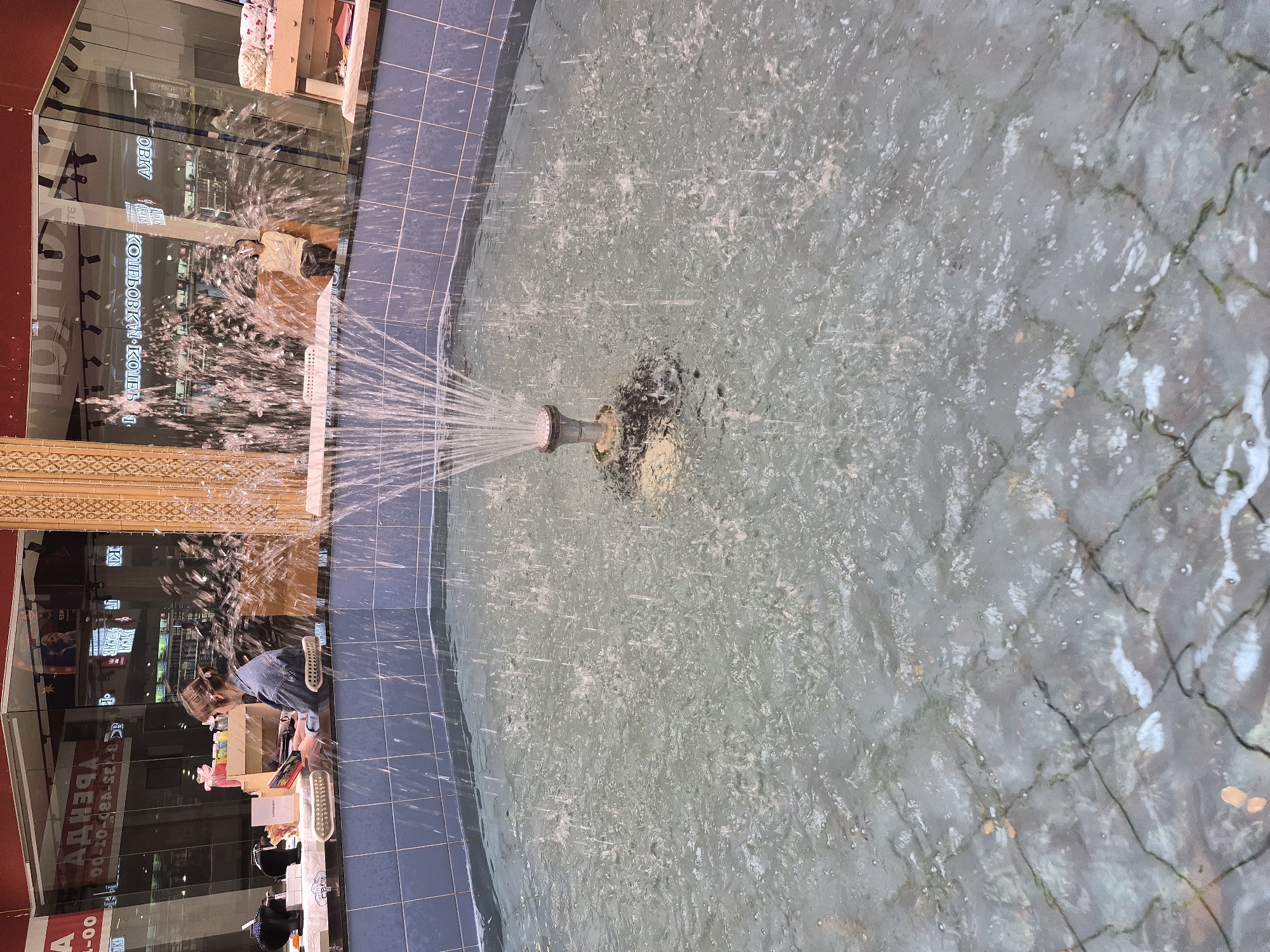 An eye-level, rotated, medium shot depicts a fountain with a woman standing on the edge, apparently at a retail shop location in Tobolsk, Russia. The fountain has a nozzle spraying water upwards, creating a radial pattern. The water surface is rippled and somewhat murky. The fountain is bordered by square blue tiles.
To the left of the fountain, a woman with her hair pulled back stands with a box of merchandise. There are several items on the shelf in front of her. She is angled toward the shop behind her. The shop's signage is in Russian. Behind the shelf are retail displays. A partial reflection is visible in the storefront windows.
