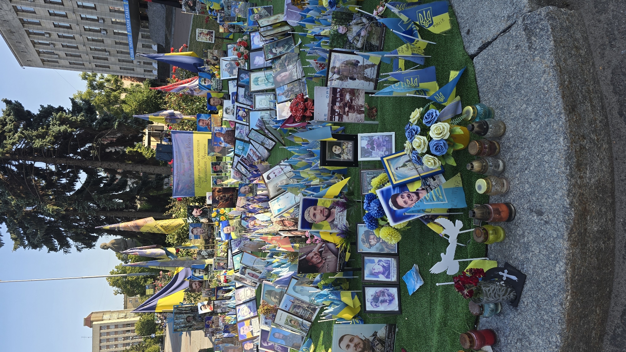 The image depicts a memorial display in the city of Zhytomyr, Ukraine, likely dedicated to fallen soldiers. The scene is set outdoors, with a large building and trees visible in the background, suggesting a public square or park. The foreground features a dense arrangement of framed photographs of individuals, predominantly young men in military attire. These photographs are interspersed with numerous small Ukrainian flags and larger Ukrainian flags are also prominently displayed. Bouquets of artificial flowers, both blue and yellow, and some real red flowers are placed amongst the photos. Several glass candle holders, some with lit candles, are arranged on a stone ledge in the lower foreground, alongside what appear to be small memorial markers. The overall impression is one of a solemn tribute.