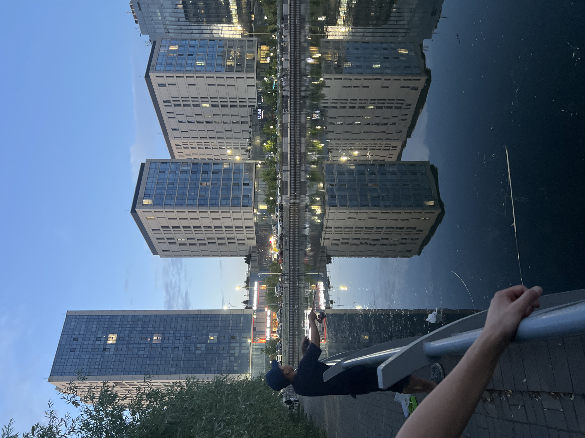 In Astana, Kazakhstan, at dusk, a person stands on a brick-paved promenade, leaning against a metal railing, fishing in a body of water. The person is wearing a dark t-shirt and a dark baseball cap. Fishing rods are cast into the water. Across the water, a row of modern buildings with glass facades and illuminated windows reflects in the still water. A few people are visible further down the promenade. The sky is a soft blue, transitioning into twilight.