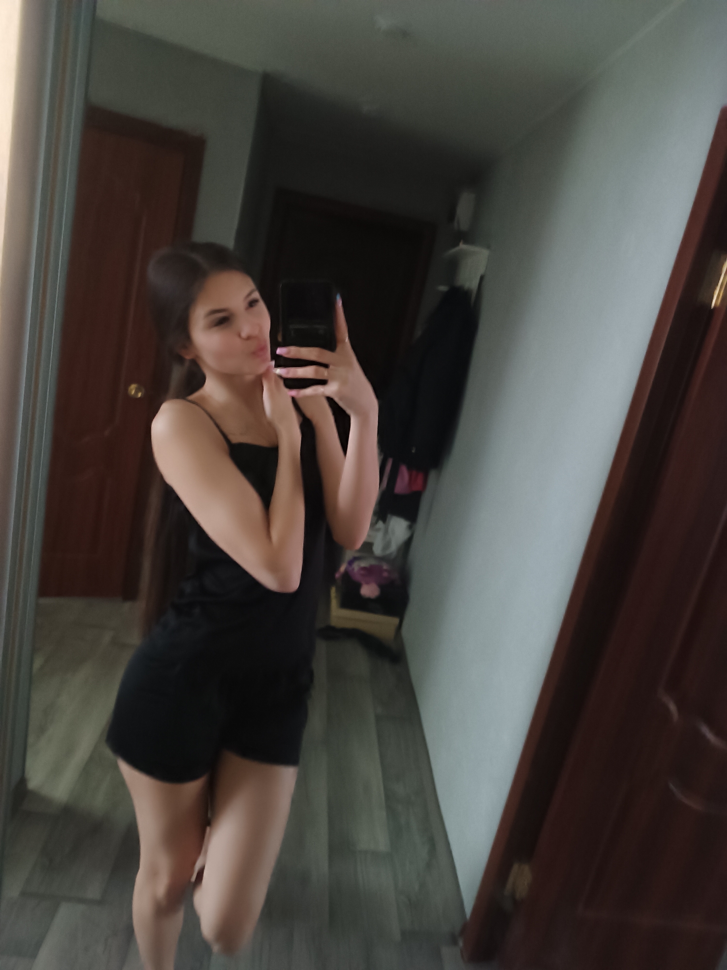 A young woman is captured in a mirror selfie, standing in what appears to be an indoor room. She has long, dark hair and is wearing a black spaghetti strap top and black shorts. She is holding a dark-cased smartphone in her right hand to take the photo, while her left hand is raised towards her neck. Her lips are puckered in a "kissing face" expression. Her fingernails are painted a light color, possibly pink or purple, and she wears a ring on her right hand.
The background, reflected in the mirror, shows a simple interior. To the left of the woman's reflection is a dark wooden door. Another dark wooden door is visible further to the right, partially open to reveal a darker space within. The walls are a light, neutral color. The floor has a light-colored wood or laminate pattern, possibly chevron or plank style. Behind the woman's reflection, to the right, a clothes rack or hanger with dark clothing and some colorful items (possibly pink/purple fabric) on a lower shelf or surface can be seen. The lighting suggests an indoor setting, likely daytime or well-lit by artificial lights. There is no visible text in the image.