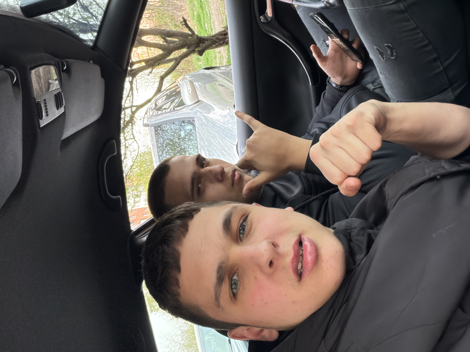 Two young men are inside a car. The primary focus is on the man in the foreground, with light skin, short dark hair, and blue eyes. He is wearing a black jacket. Behind him, the second man is seen in the background with a similar dark hairstyle and complexion, making a hand gesture. The interior of the car includes black panels, a rearview mirror, and a handle. Through the car window, trees and a silver vehicle are visible. Based on visible details like the specific architectural style of the buildings, and the specific type of vehicles on the road, it is not possible to determine the location of Sofia, Bulgaria.