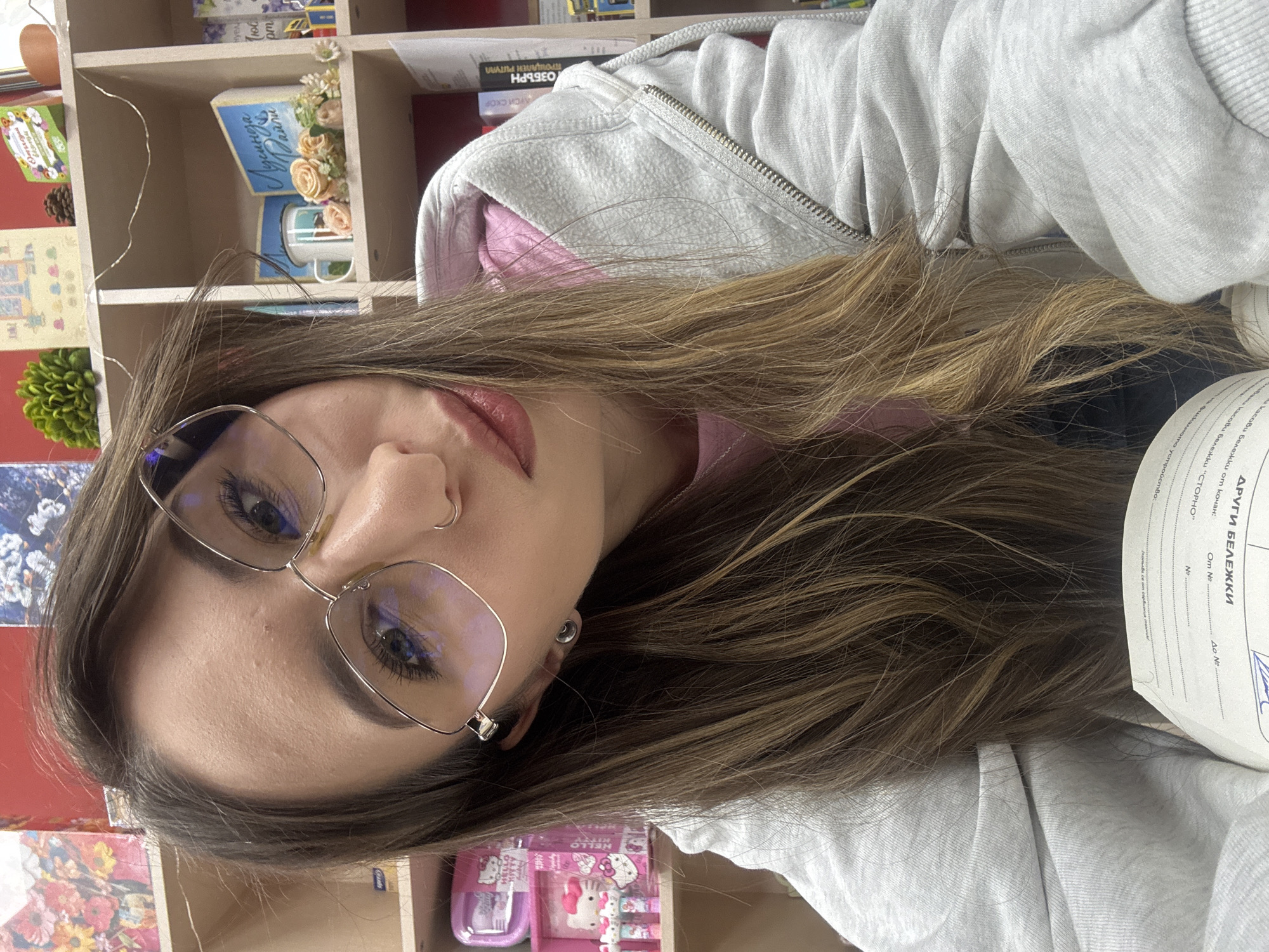 A woman with long brown hair is wearing glasses and a gray hoodie. She is near a bookshelf with various items, including books and decorations. There is a document in view with Cyrillic writing (likely Bulgarian), possibly indicating a location in Bulgaria. Based on the text "УСИ СКОР ОЗБЪРН," it could be referencing "Училищен Спортен Клуб ОЗБЪРН," which translates to "School Sports Club OZBARN." It is difficult to conclusively place the exact location in Veliko Tarnovo from just the image. The background suggests an indoor setting, perhaps a home or library.