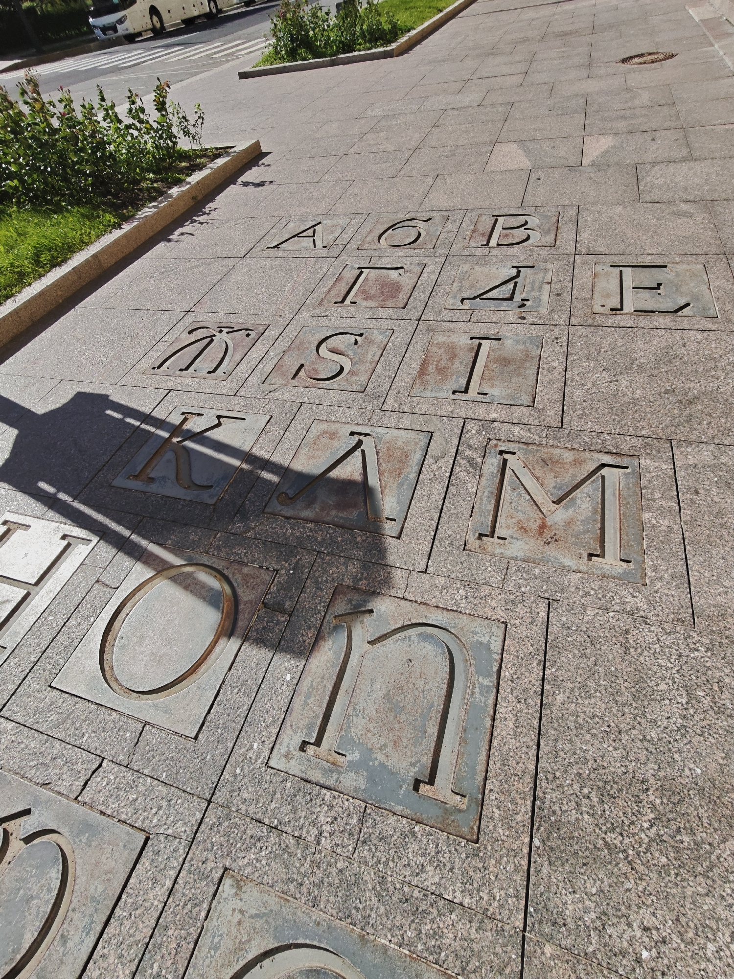 The image depicts a stone pavement with inlaid letters and numbers, likely part of an outdoor art installation or interactive display in Saint Petersburg, Russia. The pavement is made of granite slabs. Several squares are visible, each containing a Cyrillic letter or number cutout, some appearing rusted or weathered. These include letters like 'А', 'Б', 'В', 'Е', 'Ж', 'И', 'К', 'Л', 'М', 'Н', 'О', 'С', and numbers like '6', '4'. A shadow of an unseen object, possibly a lamppost or a structure, falls across the pavement, obscuring some of the letters. In the background, a bus is visible on a road, and green bushes and grass are present along the edge of the pavement. The overall setting suggests a public space in Saint Petersburg.