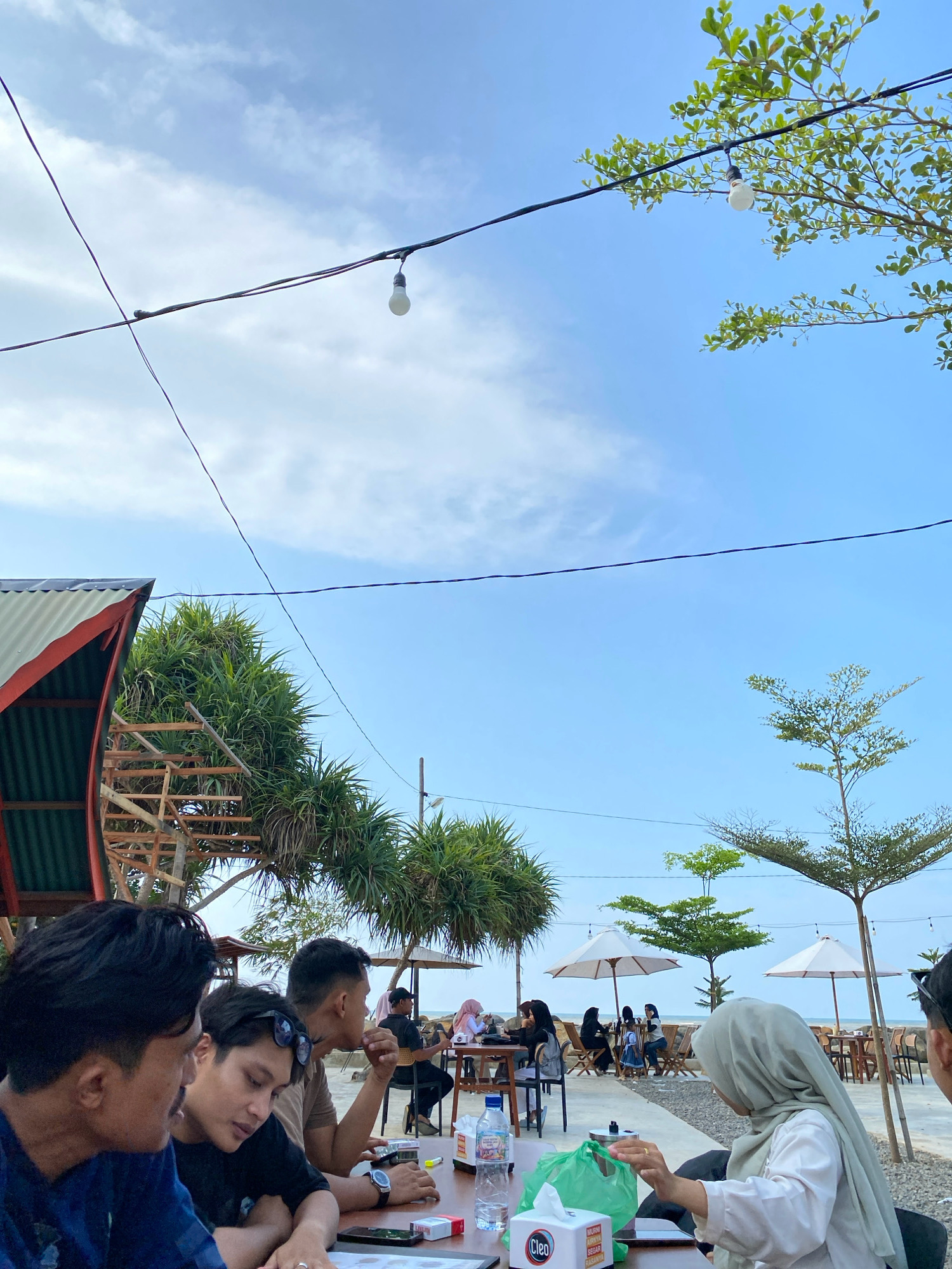 In Lancok, Indonesia, a group of people are dining outdoors at tables set up on a gravel area near a body of water, likely the ocean, under a clear sky with scattered clouds. In the foreground, three men are seated at a table, engaged in conversation. One man, wearing a blue shirt, is partially visible from the side. Next to him, a man in a black shirt and sunglasses leans forward, looking down. Across from them, another man in a brown shirt is seen from the side, holding his hand near his mouth. On the table are a smartphone, a pack of cigarettes, a bottle of water, and a green plastic bag. Further back, other diners are seated at tables, some under umbrellas. The scene is decorated with string lights and foliage, including palm-like trees and a larger tree with sparse green leaves in the background. A building with a corrugated metal roof is visible on the left side of the image.