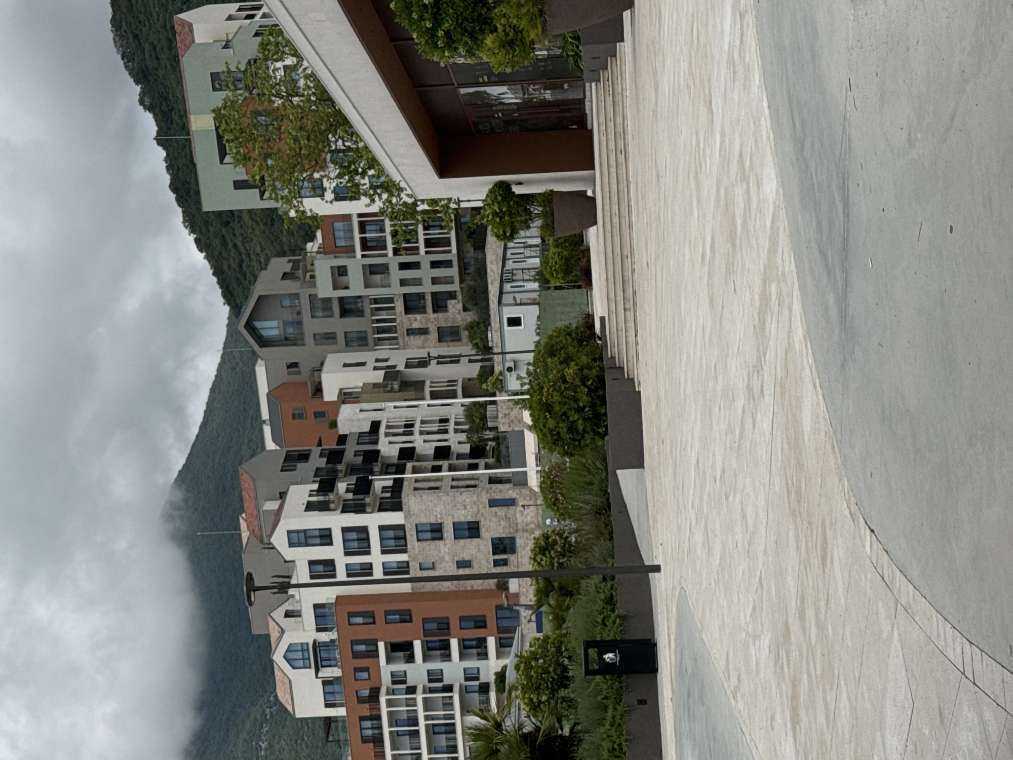 The image shows a view of a modern residential complex in Tivat, Montenegro, situated near a mountain shrouded in mist. The complex features multiple buildings with a mix of brown, white, and gray facades and numerous windows. The architecture appears contemporary, with some buildings having peaked roofs. A paved walkway occupies the right side of the frame, composed of large light-colored slabs with curved edges. The walkway is partially lined with low shrubbery and a black trash receptacle is visible near the bottom. The background is dominated by a forested mountain partially obscured by fog or clouds, lending a dramatic and somewhat overcast atmosphere to the scene.