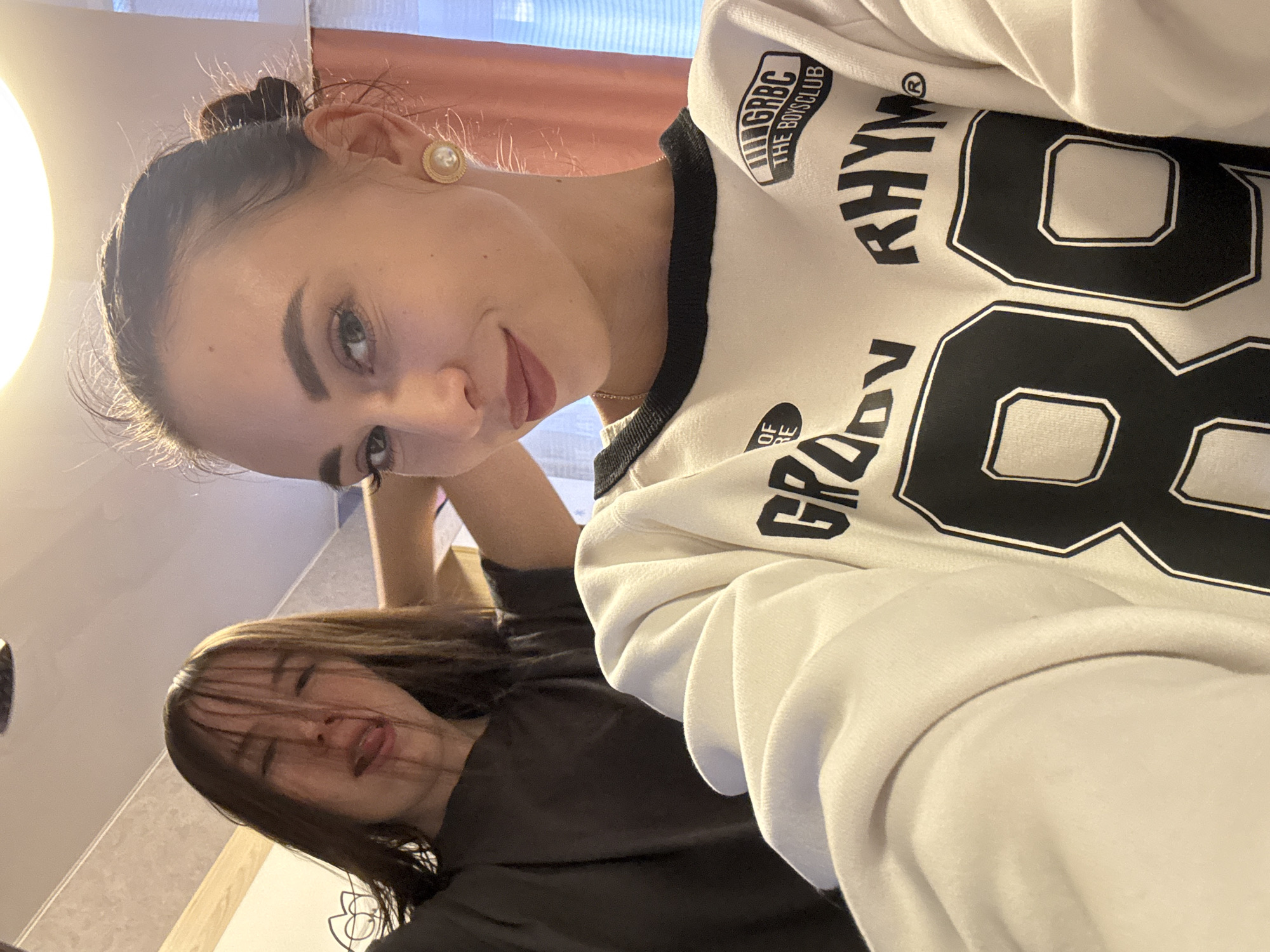 This indoor photo, taken in Chaikovsky, Russia, features two young women in a casual setting. The image is captured from a slightly low, tilted angle, with a bright light source on the left side illuminating the scene.
The primary subject in the foreground is a woman looking directly at the camera with a gentle smile. She has dark hair styled in a bun and is wearing a light-colored (likely white or cream) sweatshirt with black accents. A notable detail is the gold-rimmed earring with a pearl-like center on her right earlobe. Around her neck, a delicate gold chain is visible.
The white sweatshirt features several pieces of black text and numbers. Clearly visible are "GRBC THE BOYSCLUB" as a logo on the upper left chest area, and large stylized numbers "88" prominently displayed. Parts of the words "RHYM" and "GRODY" are also discernible on the sweatshirt.
Behind her, partially obscured by the foreground subject, is another woman. Her dark hair falls over her face, obscuring her eyes, suggesting a relaxed or playful pose. She is wearing a dark-colored top. Both subjects appear to be in a comfortable, informal environment, possibly taking a selfie or a casual portrait. The overall mood of the image is relaxed and friendly.