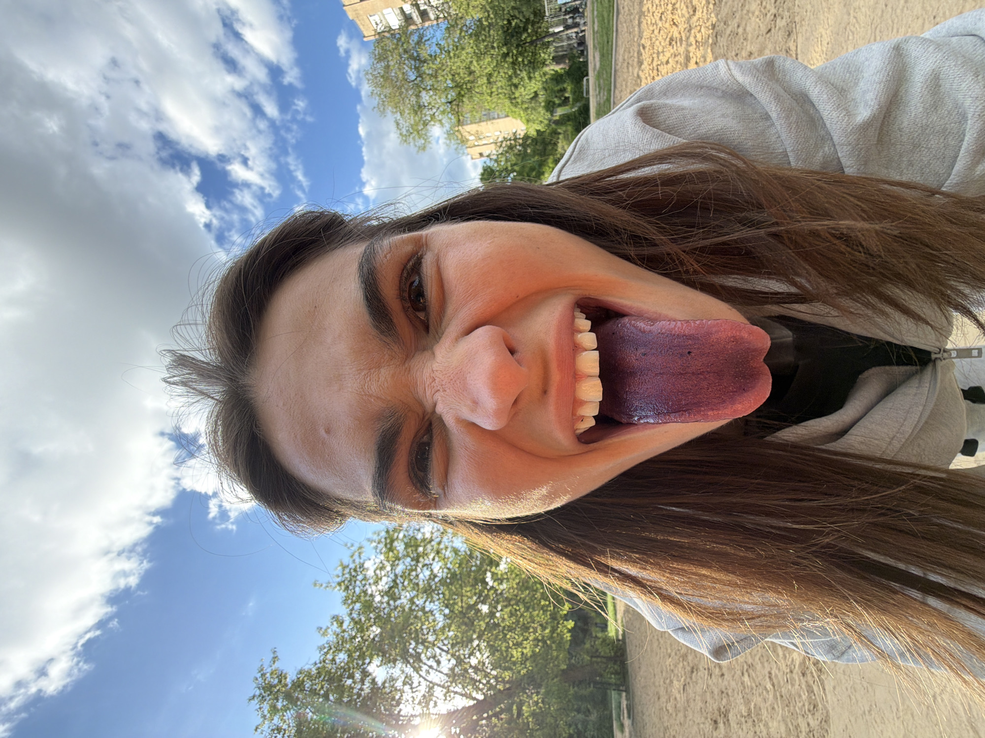 A woman is the subject of a selfie, her face angled upward to capture the sky and trees in the background. Her features are exaggerated due to the proximity to the camera; her eyes are squinted and her nose is wide. She is wearing a grey hooded sweatshirt. Her mouth is open, revealing a bluish-purple tongue and set of teeth. A glimpse of blue sky with scattered clouds is visible above her head. Trees are also in the background, with sunlight shining through the leaves. The ground appears sandy or earthy. The overall impression suggests an outdoor setting, potentially a park, within a city. While the specific location in Moscow, Russia, cannot be determined from the image, the presence of green spaces and residential buildings align with typical urban landscapes.