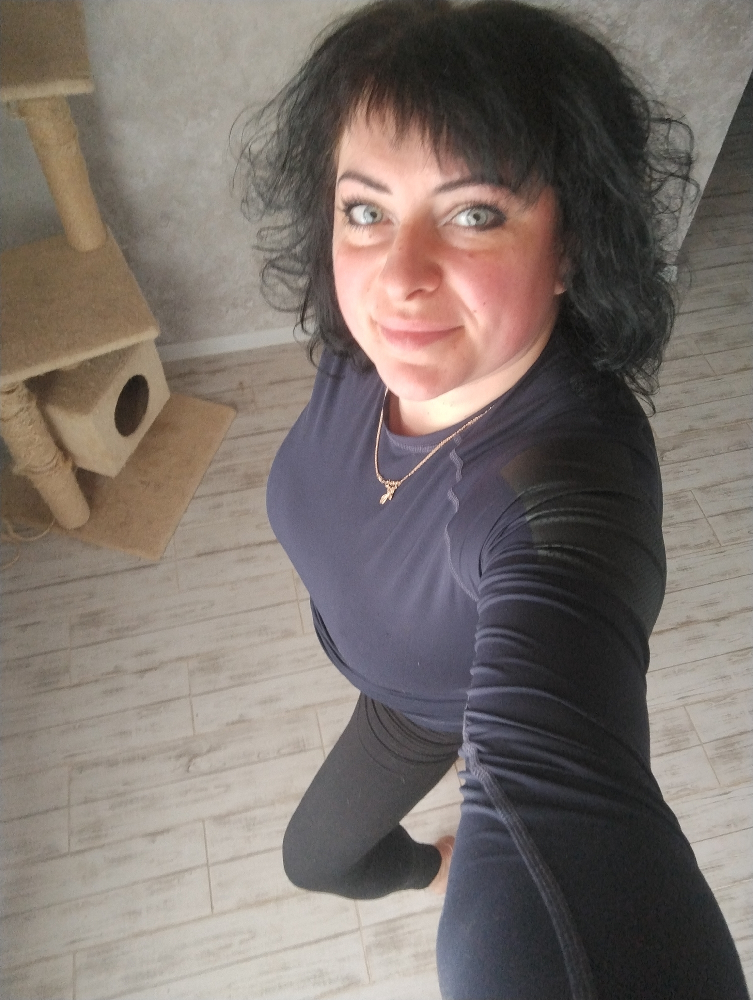 The image is a selfie taken from above, featuring a woman with fair skin, blue-green eyes, and dark, wavy hair with bangs. She is wearing a dark-colored, long-sleeved athletic shirt and dark leggings. A gold necklace with a small pendant is visible around her neck.
In the background, there is a light-colored wooden floor and a light gray wall. A cat tree with multiple levels and a small enclosure is visible to the left of the woman.
Given the request, it is assumed the image was taken in Grishanki, Russia, although there is no way to confirm this based on the visual information alone. The specific location within Grishanki is not discernible. The scene appears to be indoors, possibly in a residential setting.