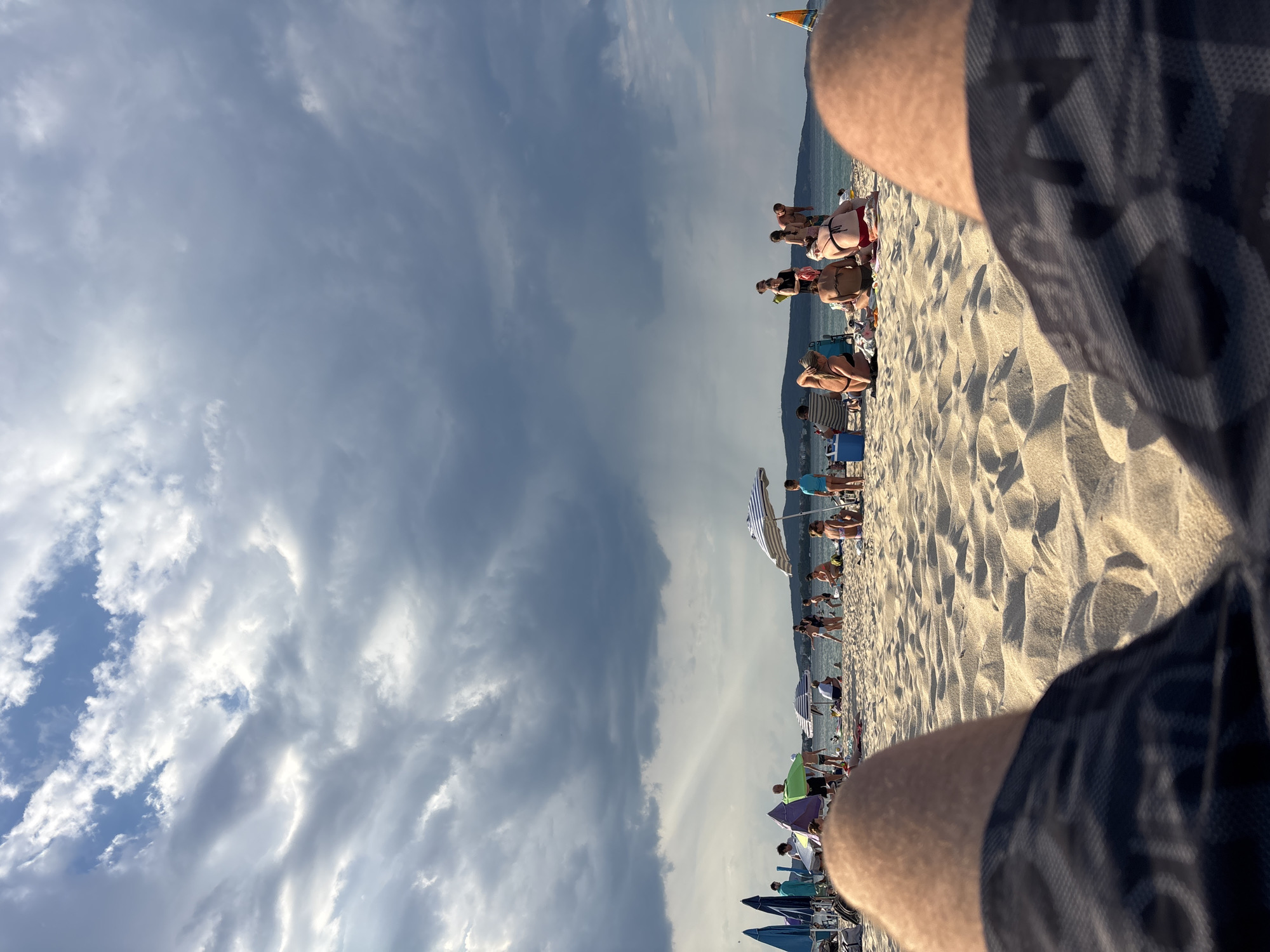 The image depicts a crowded beach in Kiten, Bulgaria, under a cloudy sky. The perspective is from someone seated on the sand, looking towards the sea. In the foreground, the legs of the seated person are visible, wearing patterned shorts. The sand is textured with ripples and indentations.
Further back, numerous people are scattered across the beach, engaged in various activities. Some are sitting on towels or chairs, others are standing or walking. Several beach umbrellas, some striped and others solid blue, are open, providing shade. A cooler is visible near some beachgoers. In the background, the sea is calm, and a distant coastline with hills is visible. The sky is dominated by dramatic, dark clouds, with patches of lighter sky and sunlight breaking through in some areas. The overall atmosphere suggests a lively beach day, potentially with impending weather changes due to the cloudy sky.