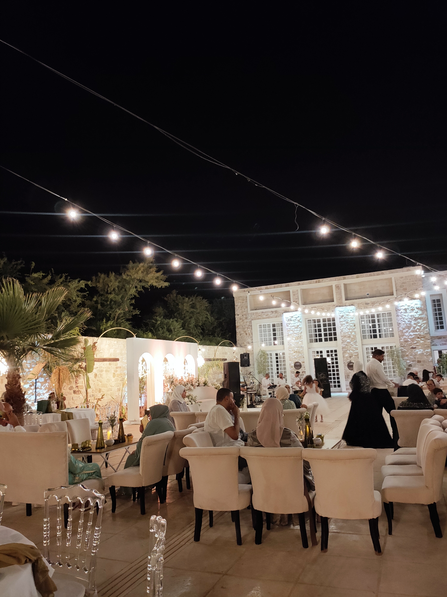 The image depicts an outdoor evening event at the "La Casbah" venue in Cebalat, Tunisia. A group of people are seated at tables, with some engaged in conversation while others appear to be observing an ongoing activity. In the background, a building with large windows and stone walls is illuminated, suggesting a reception or celebration. Strings of lights are strung across the scene, providing illumination. A band or musical ensemble is visible performing in front of the building. Some individuals are dressed in formal attire, and the overall atmosphere suggests a festive gathering. A young girl in a white dress is also present, potentially participating in a wedding or similar ceremony. The venue appears to be a spacious outdoor area with palm trees and lush greenery visible.