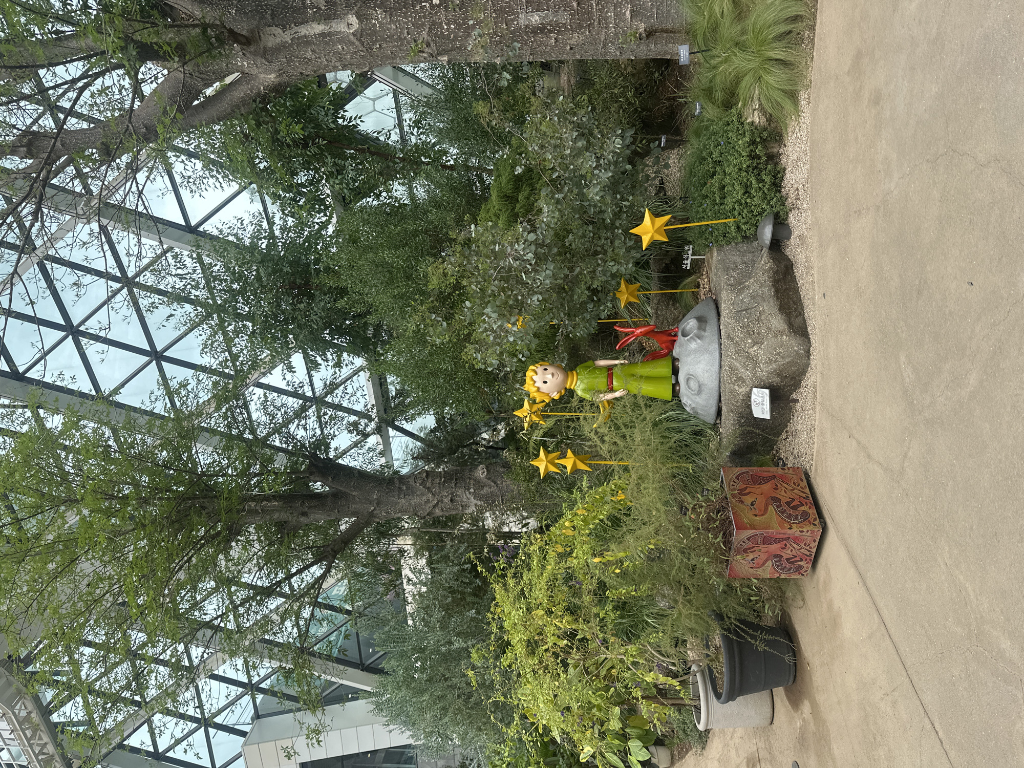 The image shows a decorative display of "The Little Prince" statue in a botanical garden, likely within the city of Seoul, South Korea. The statue depicts the Little Prince in his characteristic green outfit, standing on a large rock that resembles a moon. Beside him is a small red fox. Yellow star decorations are strategically placed around the display. The background features lush greenery, trees, and a modern glass-domed structure with a geometric pattern, characteristic of many urban botanical gardens. Two large potted plants and a decorative planter with an intricate design are visible to the left of the display, on a concrete pathway. The lighting suggests it is daytime, with diffused light filtering through the glass dome.