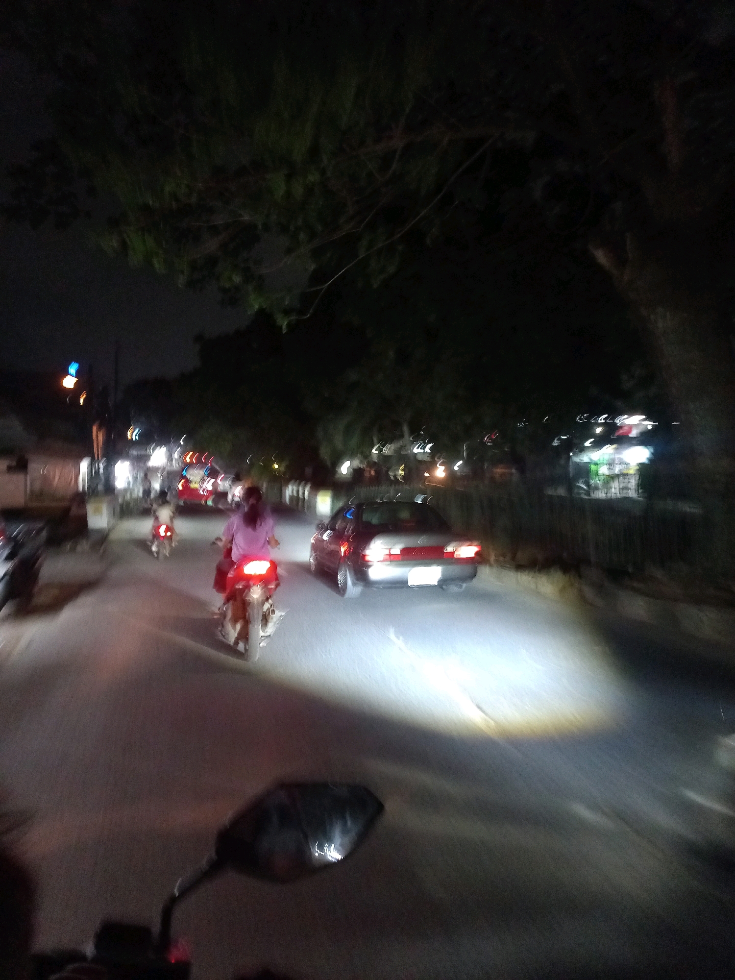 The image depicts a nighttime street scene in Depok, Indonesia, characterized by motion blur and low light conditions. The perspective appears to be from a moving vehicle, likely a motorcycle, with a side mirror visible in the foreground. Several motorcycles are present, with one rider wearing a pink shirt in the mid-ground, illuminated by headlights. A silver sedan is also visible on the road, its headlights casting a bright beam. The street is lined with trees, and there are some lights from buildings in the background, suggesting an urban or suburban environment. The overall impression is of a busy street at night, with traffic and ambient illumination.