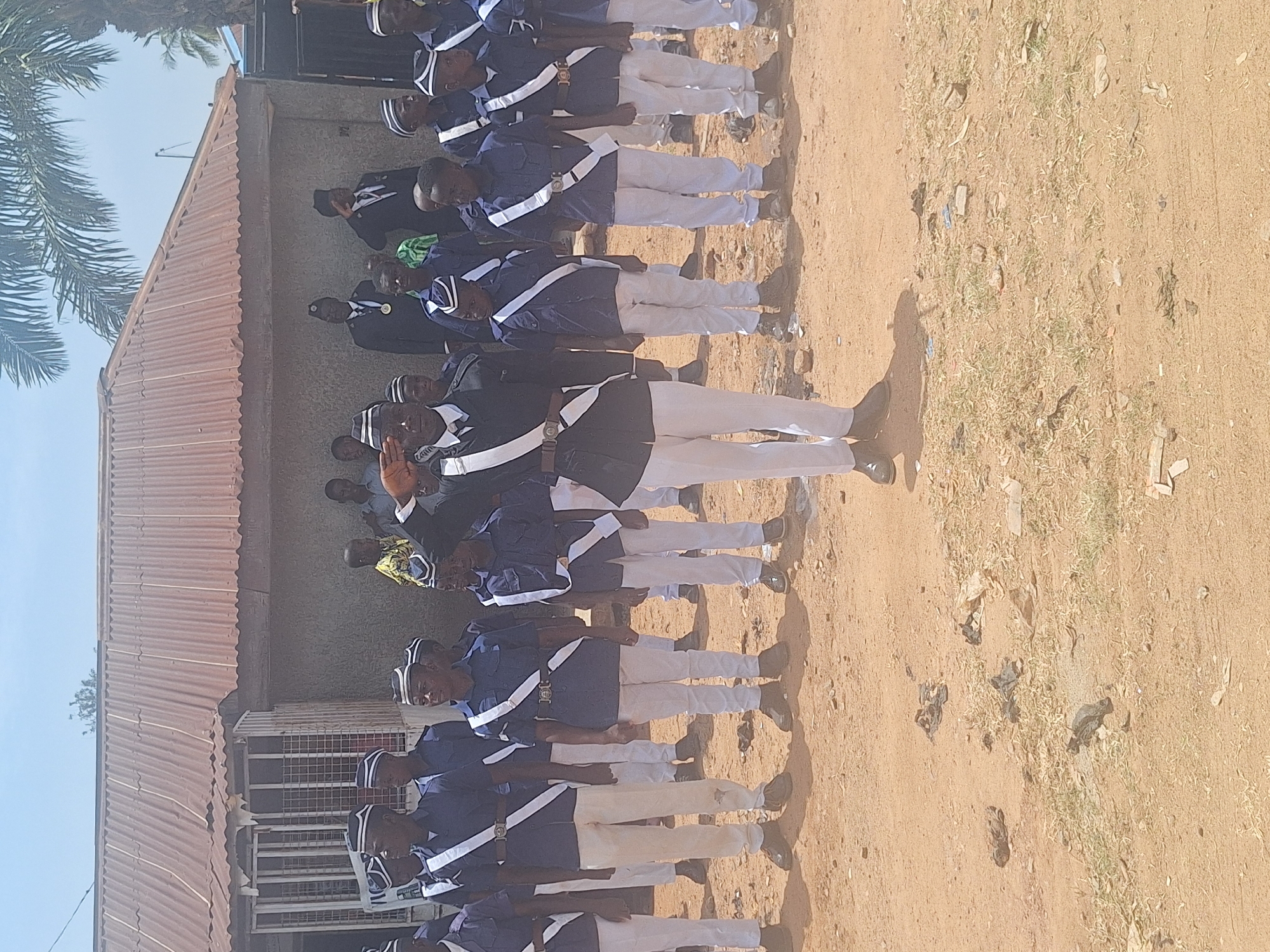 In Sabon Tasha, Nigeria, a group of uniformed individuals, dressed in navy blue shirts, white pants, and white hats, stand in formation on a dirt surface. A building with a corrugated metal roof is visible in the background. One individual, wearing a black suit jacket over his uniform, salutes. Some people stand behind him. The ground is littered with debris and sparse vegetation. A tree with distinctive foliage is visible on the left side of the frame.