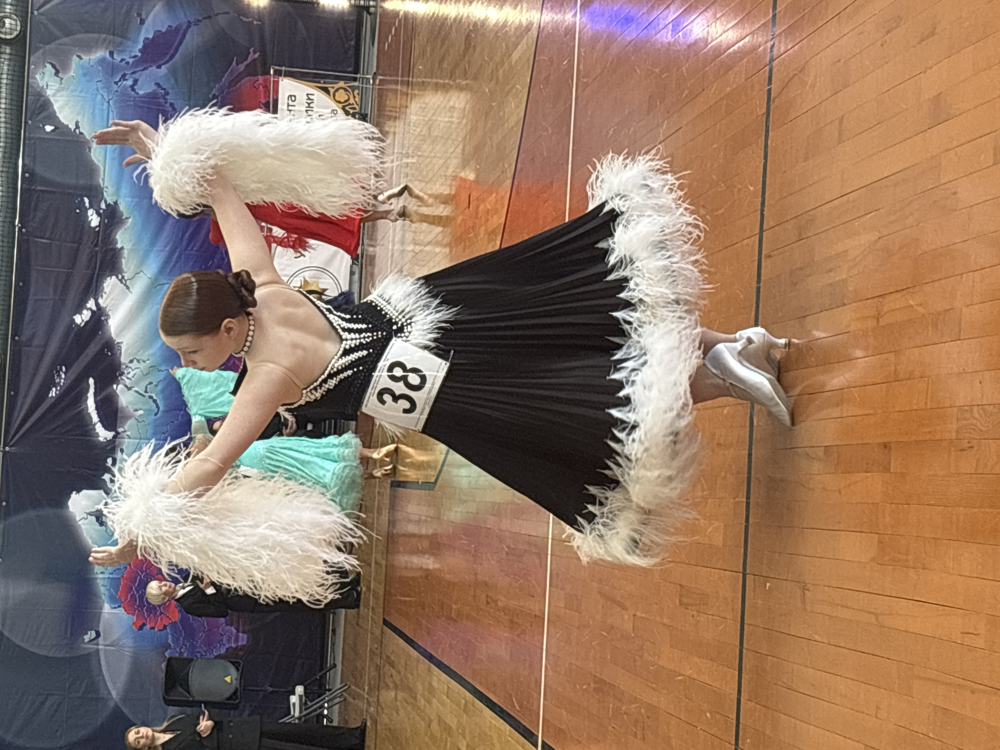 A woman in a black and white ballroom dancing dress with feather boas is competing in a ballroom dance competition. The number "38" is pinned to her dress. The competition appears to be taking place on a wood dance floor in a gymnasium. A backdrop with world map graphics is visible behind the dancers. The location is potentially in Izhevsk, Russia, based on possible context clues in the image, such as signage on the backdrop. Other dancers are visible in the periphery.