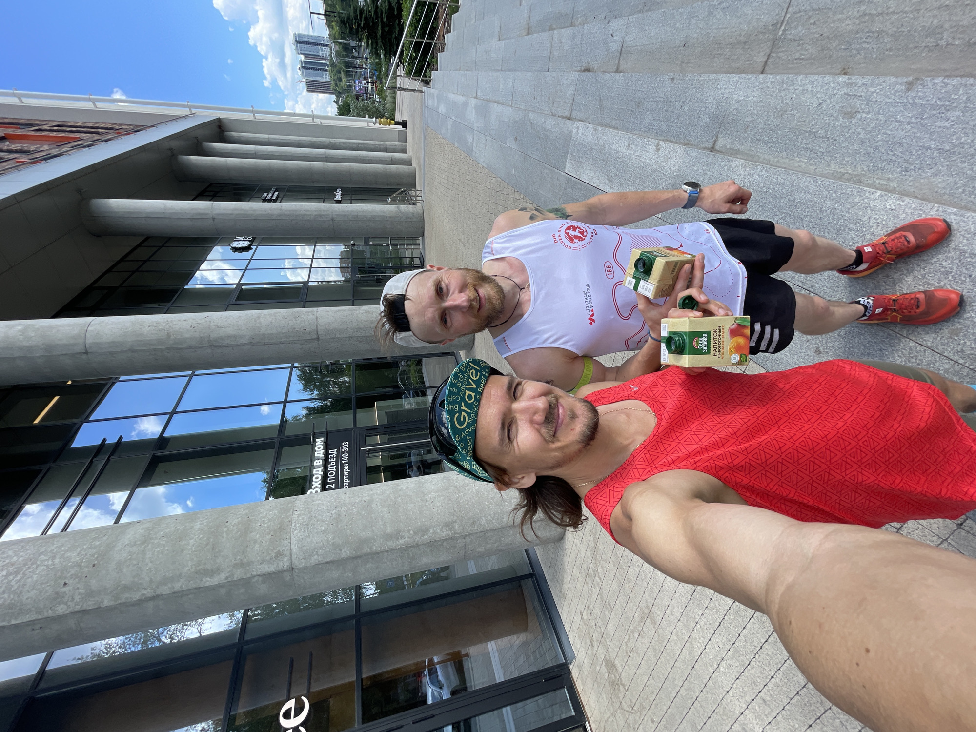 Two men, appearing to be runners, are standing on a sidewalk in front of a modern building with a glass facade. The man on the left, wearing a red tank top and a green cap, is taking a selfie. He has long, dark hair and is smiling. The man on the right, wearing a white tank top and a grey baseball cap, is holding two small juice boxes, one open and one closed. He also has a beard and is smiling. Both men are wearing athletic shorts and running shoes. The building behind them features large concrete pillars and reflective glass windows. In the background, there are trees, a blue sky with scattered clouds, and distant city buildings. The overall scene suggests an outdoor urban environment, possibly after a run.