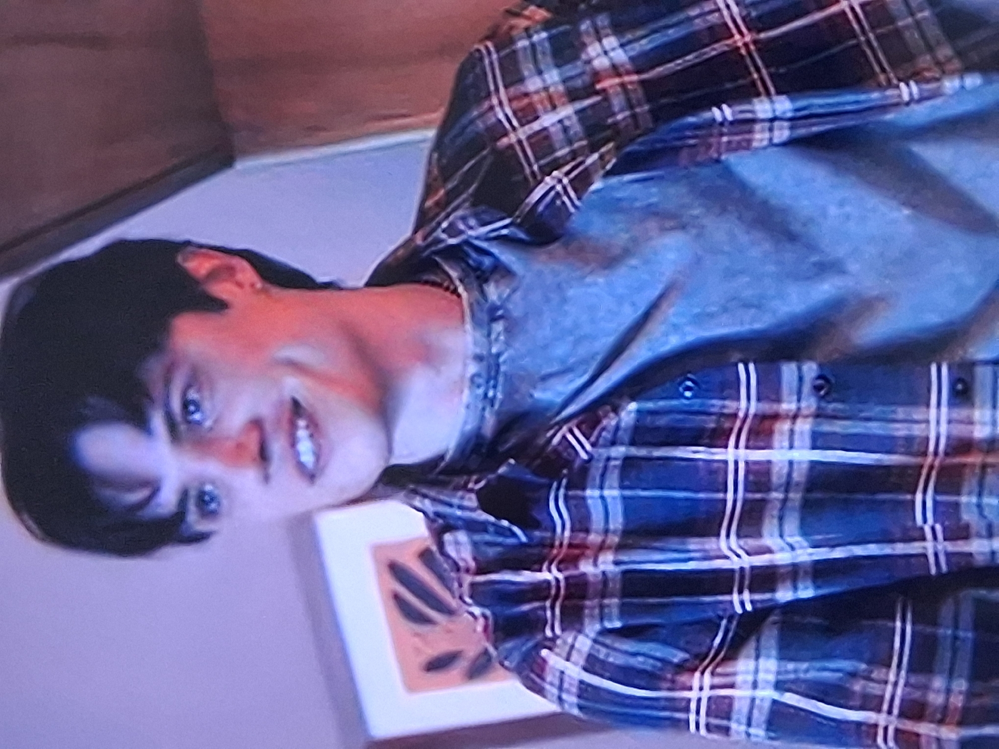 A young Caucasian male with dark hair is shown from the chest up. He is wearing a grey t-shirt and an open, dark blue and red plaid shirt over it. His eyes are blue, and he has dimples. He is smiling, and his teeth are visible. He has an earring in his left ear. The background is blurred but appears to be an indoor setting with a picture on the wall. There is no indication of the city of Semey, Kazakhstan, in the image.