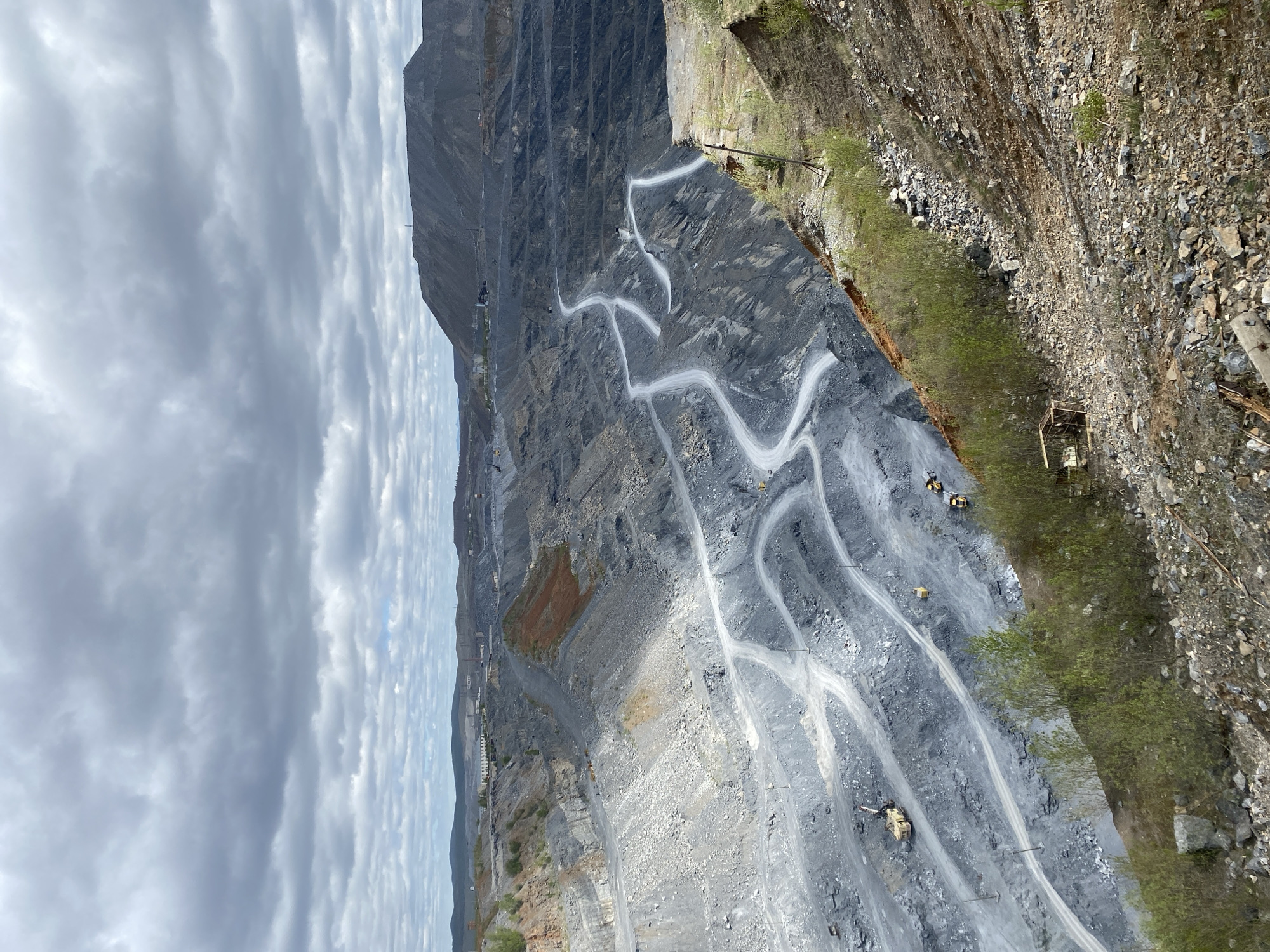 The image depicts a large open-pit mine located in Satka, Russia. The mine features terraced layers with winding roads traversing the levels. Several pieces of heavy machinery, possibly excavators or trucks, are visible on the roads. The pit walls are primarily gray in color with some areas of orange and brown discoloration. Overhanging the mine is a cliff covered in loose stones and some scrub vegetation. The sky is filled with gray clouds.