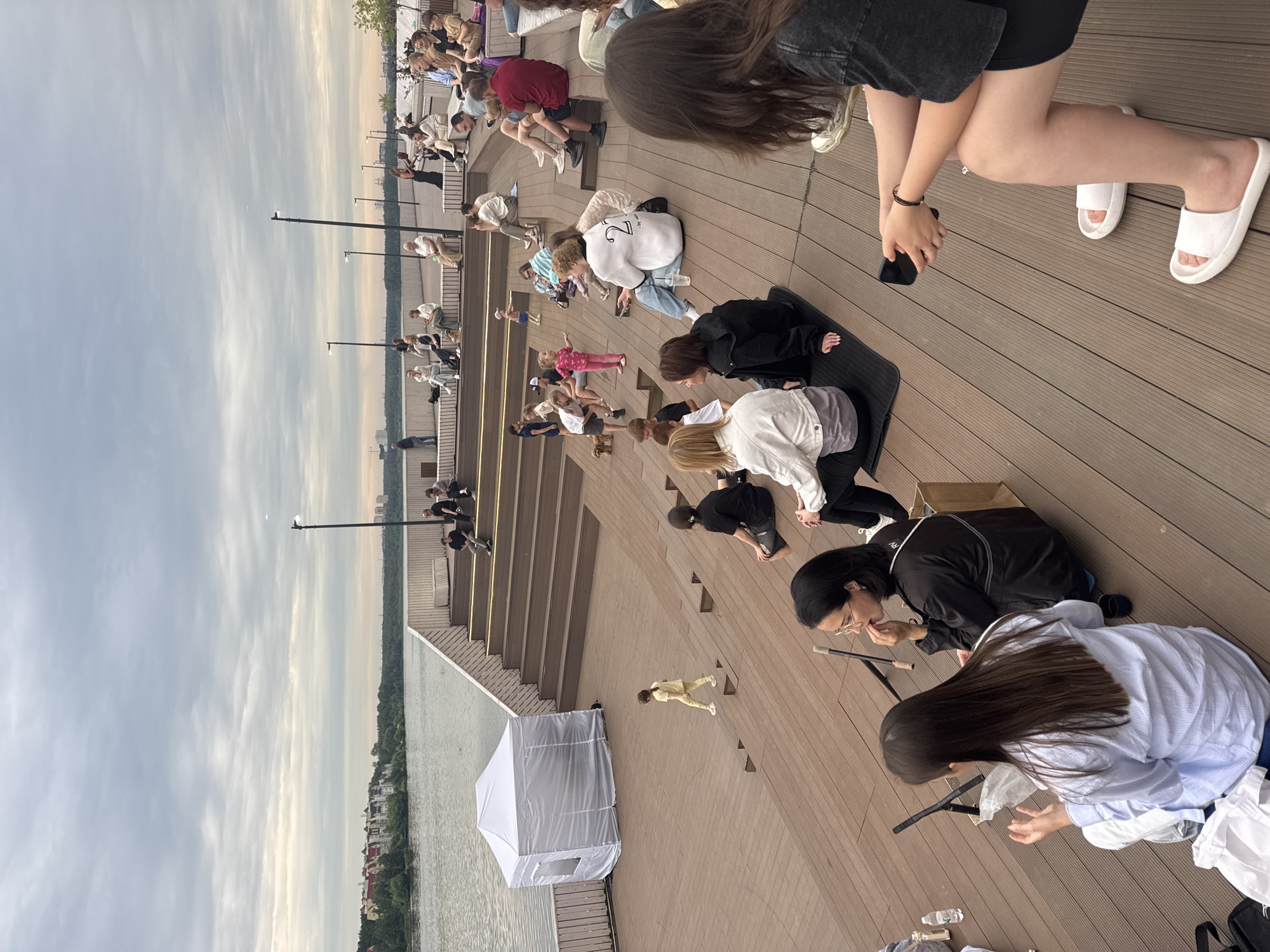 This image captures an outdoor event at the Yekaterinburg City Beach, also known as "New Timur," situated on the Iset River embankment. The scene depicts a crowd of people gathered on a tiered wooden seating area, resembling an amphitheater. The event appears to be a performance or lecture, as indicated by the presence of a small white tent and a few individuals in the foreground who seem to be observing. The sky is overcast, suggesting a cloudy day. In the background, a body of water, presumably the Iset River, is visible, with buildings and trees lining the opposite bank. The wooden structure extends down to the water's edge, creating a modern urban recreational space.