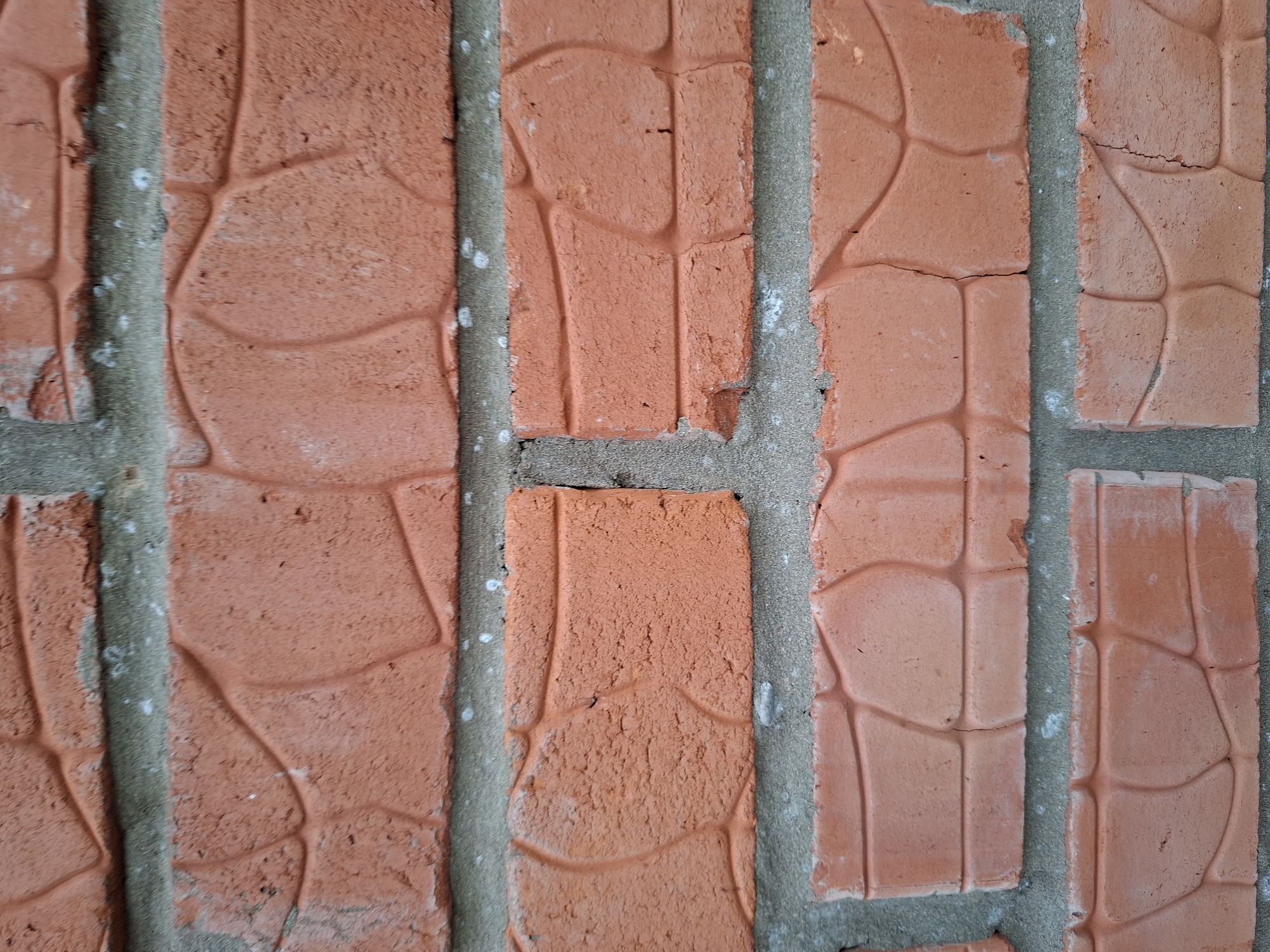 This is a close-up, eye-level, still, medium shot of a brick wall. The wall is made of horizontally laid, rectangular, orange-red bricks with a textured surface resembling stone. The bricks are laid with grey mortar, and there are visible cracks and imperfections in both the bricks and the mortar. The lighting is even, and there is no indication of time of day or weather. The image does not contain any people, animals, or vehicles. The setting is a wall, but no specific landmarks or identifying features of Votkinsk, Russia are visible.