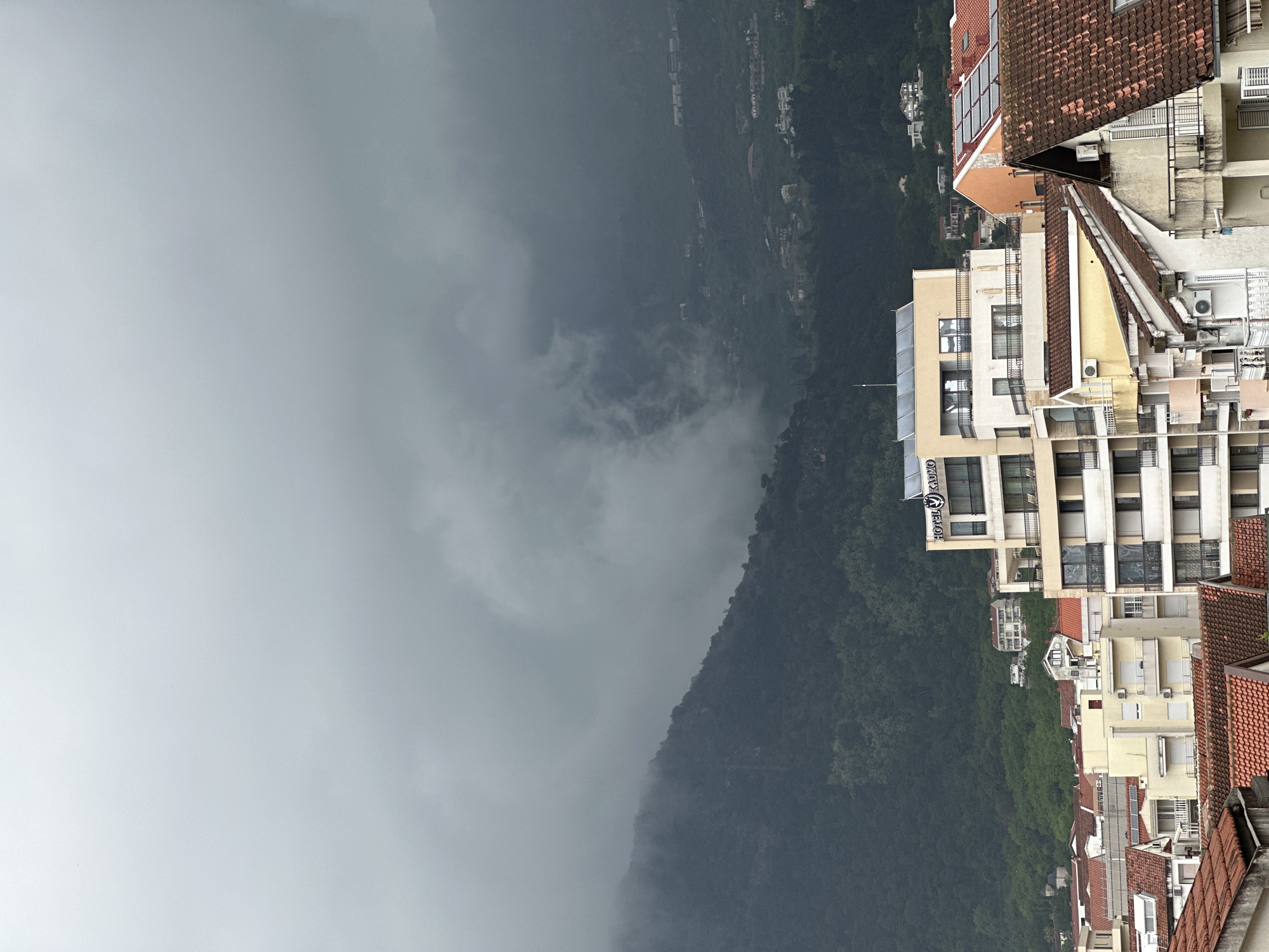 This image depicts a view from an elevated position, likely a building, overlooking a cloudy mountain landscape and part of the city of Budva, Montenegro. The lower right portion of the frame features several buildings with orange tile roofs, characteristic of Mediterranean architecture. The buildings are a mix of residential and commercial structures, with some displaying air conditioning units and window details. A portion of the facade of the "Hotel Kadmo" is visible on one of the buildings. The left side of the image is dominated by a dark, forested mountain range shrouded in low-hanging clouds or fog, creating a sense of depth and a somber atmosphere. A few scattered houses and structures can be seen dotted along the mountainside in the distance. The overall composition suggests a location where urban development meets the natural landscape. The sky appears overcast and uniformly gray.