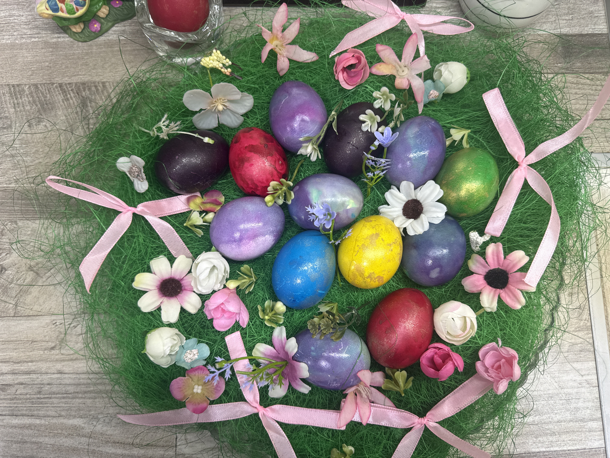 The image depicts an Easter-themed arrangement. Centered is a round wreath of green artificial grass. Scattered within the grass are approximately 15 decorated Easter eggs, various artificial flowers in shades of white, pink, and blue, and pink ribbons tied into bows. The eggs display a variety of colors including purple, red, blue, yellow, and green. The arrangement is placed on a surface with a light grey wood-grain pattern. The lighting appears bright and even. The location is not discernible from the visual elements. The presence of Easter eggs and decorations suggests a celebration or display related to the Easter holiday. No people are visible. There is no indication of Varna, Bulgaria.
