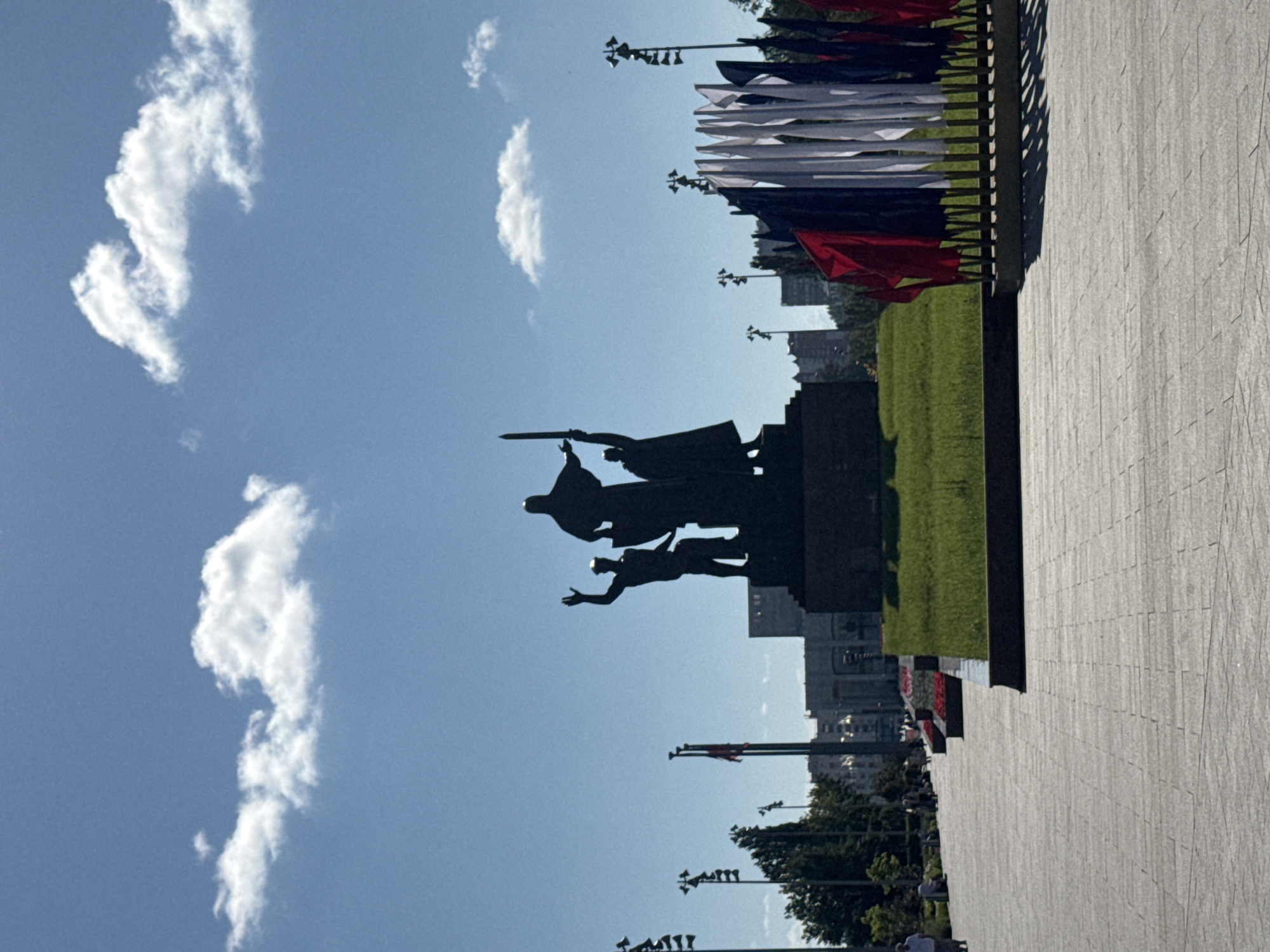 The image depicts a large, dark-colored bronze monument in a public square, silhouetted against a bright blue sky with scattered white clouds. The monument features three figures, the central and tallest figure appearing to hold a sword aloft. The figures stand on a dark, stepped pedestal. In the foreground, a paved walkway is visible, constructed with large, rectangular stone tiles. To the right of the monument, a row of red, white, and blue flags on short poles are displayed. Behind the monument, green grass and trees are present, along with buildings and streetlights in the background. The scene is illuminated by daylight, suggesting it is daytime.
This monument is the "Monument to the Fighters for Soviet Power" located in the city of Perm, Russia, on Komsomolsky Prospekt.