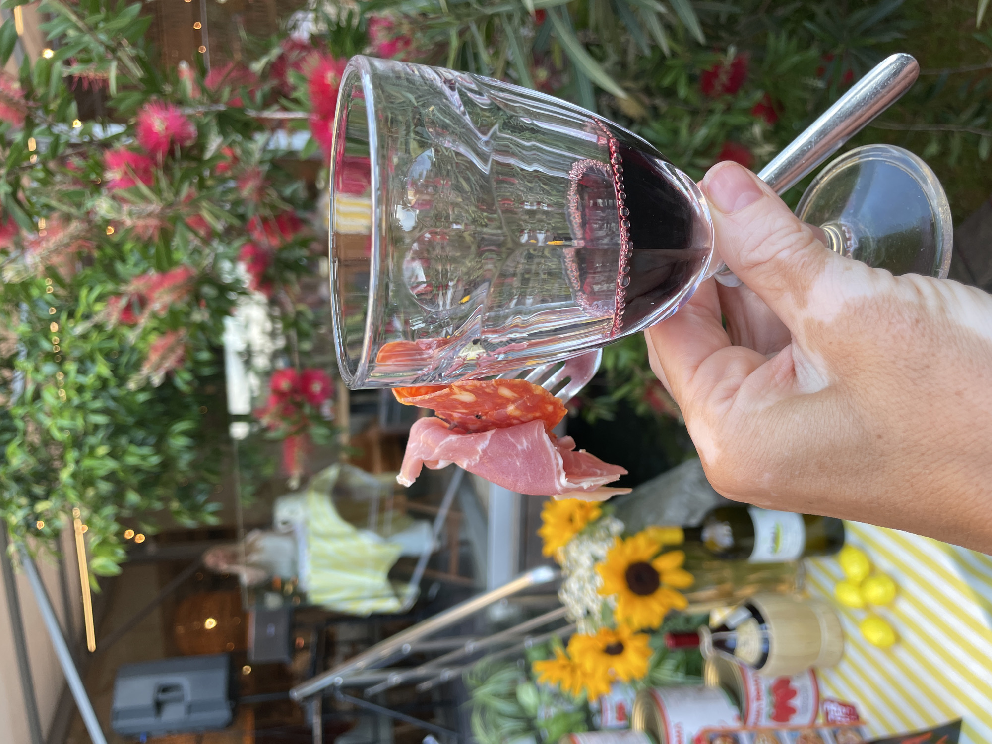 In the foreground, a person's hand with vitiligo holds a glass of red wine and a fork with two pieces of cured meat, likely prosciutto and salami. The glass is partially filled with red wine, with small bubbles visible at the surface. In the background, a table is set with sunflowers, lemons, canned goods, and bottles of wine, suggesting an outdoor market or gathering. A woman is visible in the distance, seated at a table covered with a yellow and white striped cloth, and a speaker is positioned to her left. Lush greenery and red flowers form a vibrant backdrop. The scene appears to be set in Sochi, Russia, based on the contextual information provided.