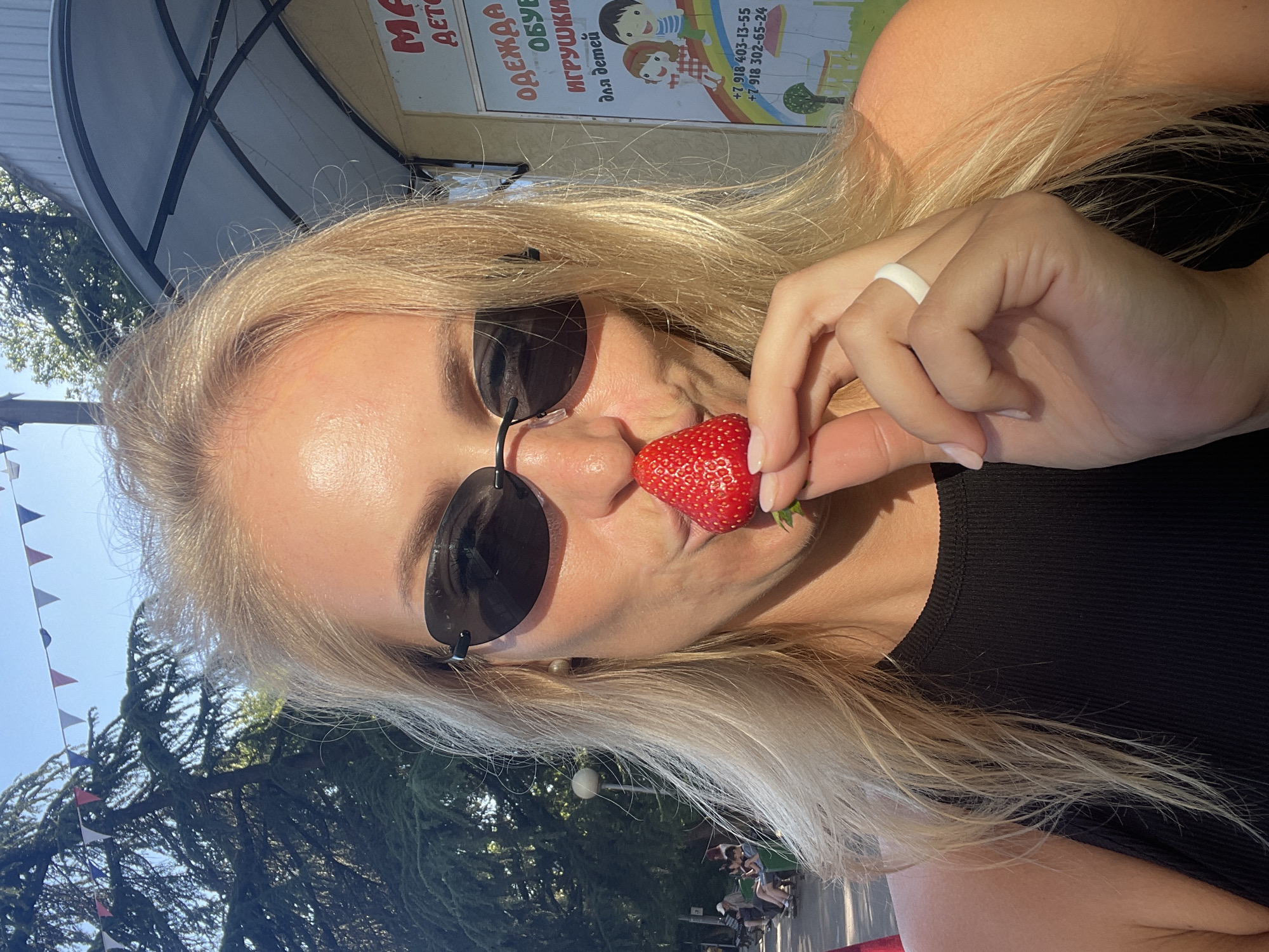 A blonde woman, wearing dark oval sunglasses and a black ribbed sleeveless top, is featured in a close-up outdoor shot. She holds a single red strawberry with her left thumb and index finger directly in front of her puckered lips, appearing poised to kiss or bite it. A white ring is visible on her left ring finger, and she has a small pearl stud earring in her right ear.
The scene takes place on a sunny day in a public park area within Sochi, Russia. In the background to the left, dense green evergreen trees are visible against a clear blue sky, strung with red, white, and blue triangular bunting flags. A lamppost and distant figures seated on park benches are partially visible. To the right, a light-colored building with a dark-framed, curved metal awning is present. A sign on the building, written in Cyrillic, advertises "ОДЕЖДА, ОБУВЬ, ИГРУШКИ для детей" (Clothes, Shoes, Toys for children) and displays two Russian phone numbers starting with +7 918, which is characteristic of the Krasnodar Krai region. Below the text, a colorful illustration depicts children, a rainbow, trees, and a small house. Strong sunlight illuminates the woman's hair, skin, and the strawberry, while casting shadows under her chin and sunglasses.