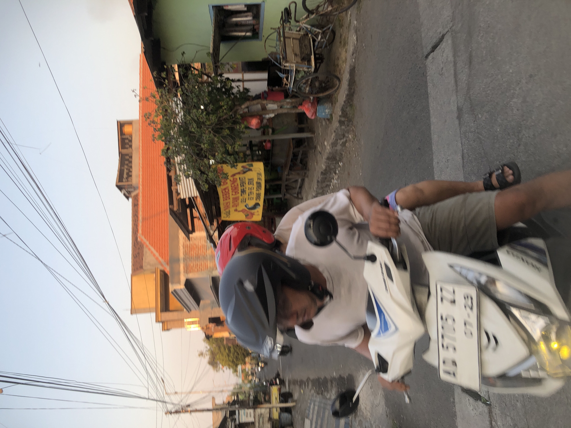 The image depicts a street scene in Tenggulunan, Indonesia, during daylight, possibly late afternoon given the lighting. Two individuals are riding a white scooter. The person in front, the apparent driver, is wearing a black helmet and a light-colored t-shirt, with their arms extended towards the handlebars. The second person, the passenger, is wearing a red helmet and is partially visible behind the driver. The scooter has a license plate that reads "AD 5705 ZZ" and "07-29".
The environment shows a paved street with a pedestrian walkway on the right. Along the walkway, there are buildings, including a green structure with a window, and a stall with a yellow banner displaying text and images of chickens. Benches are situated in front of the stall. A tree with red bags hanging from it is visible near the stall. Various objects, including a bicycle with a cart, are present on the walkway. Overhead, numerous electrical wires crisscross the sky, suspended from utility poles. In the background, more buildings with orange tile roofs are visible, along with other vehicles, including motorcycles, on the street. The overall impression is a busy, lived-in urban environment.