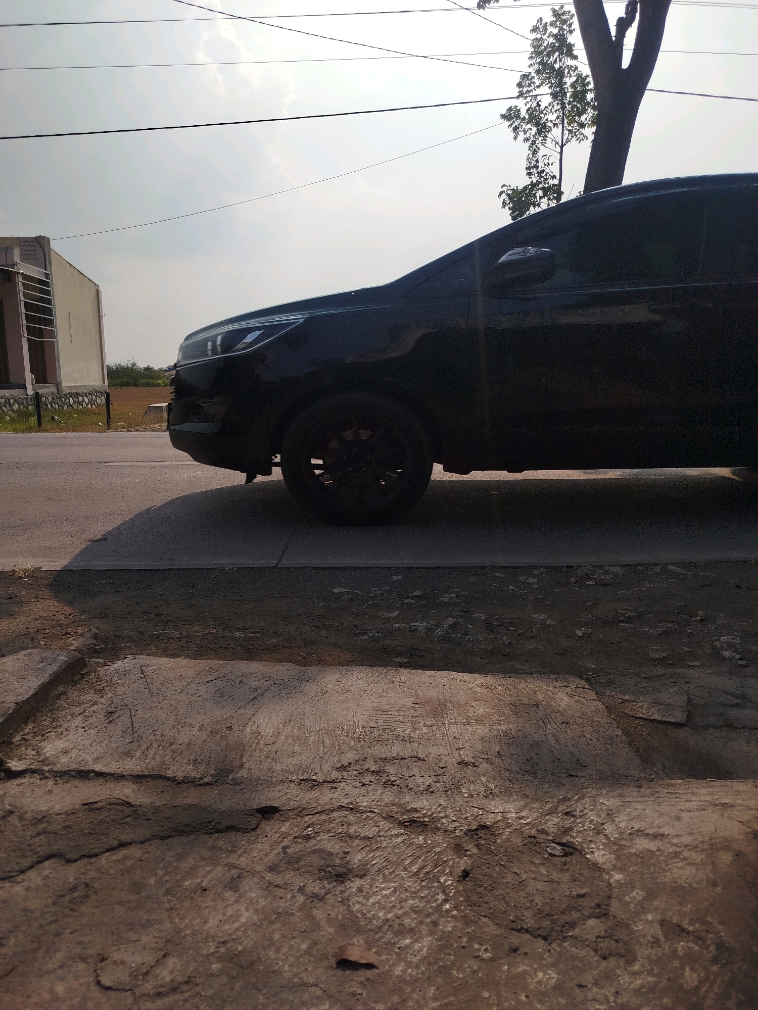 A black car is parked on the side of a road in Ngreco, Indonesia. The car is a modern SUV with tinted windows and black rims. The road appears to be paved, and there is a concrete curb in the foreground. A light-colored building with a patterned window is visible on the left side of the image, and a tree with sparse foliage is behind the car. Overhead wires are strung across the sky, which is bright and overcast. The ground is covered with dirt and some debris. The lighting suggests it is daytime, possibly late afternoon due to the long shadows cast by the car. There are no people or any obvious activity depicted in the scene.