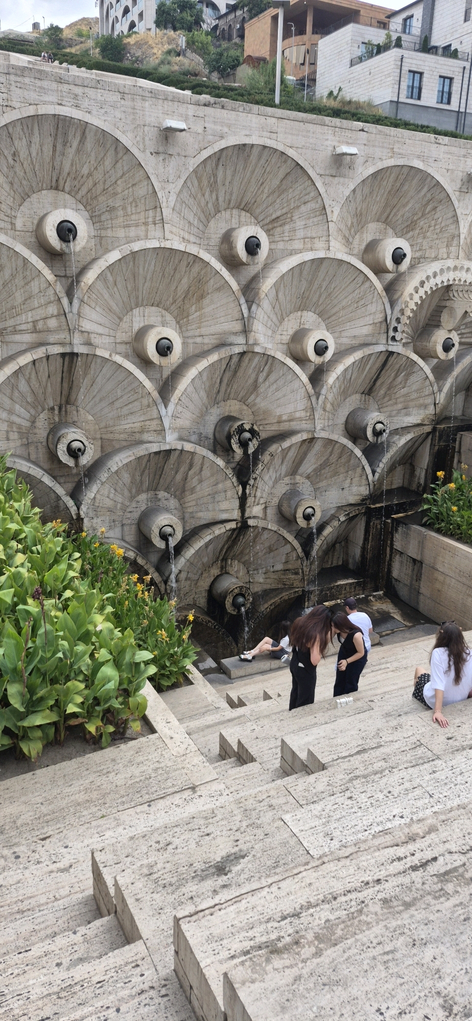 The image captures a section of the Cascade Monument in Yerevan, Armenia, a grand stairway adorned with fountains. In the foreground, a person is seated on the stone steps, facing left. Further up the steps, two individuals, one wearing black and the other white, appear to be interacting, with the person in black bending down towards the person in white. To the left, a woman is sitting on a ledge of the monument. The background features the iconic tiered structure of the Cascade, with water flowing from various spouts into pools. Lush green plants and yellow flowers border the lower part of the monument. Above the monument, modern buildings are visible on the hillside. The overall scene depicts a leisurely moment at a popular Yerevan landmark.