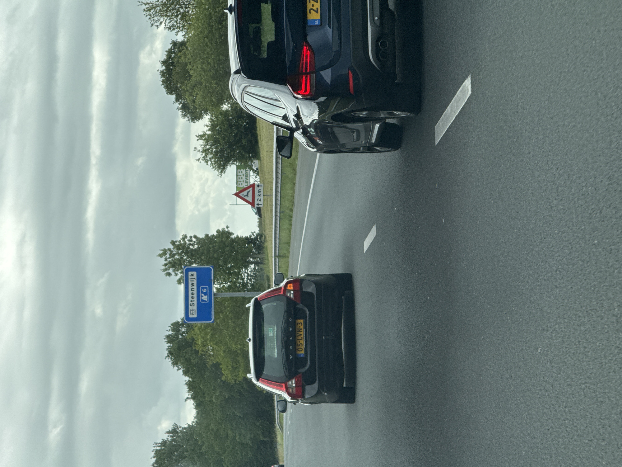 The image shows a perspective from inside a moving vehicle on a highway, looking towards the road ahead. A dark-colored Volvo station wagon, with a Dutch license plate "05-LVN-3", is in the left lane. To the right, in the right lane, is a dark blue BMW X1 SUV with a Dutch license plate "2-Z". Both vehicles are traveling away from the viewer.
In the background, a blue highway sign indicates "P+R Steenwijk" and the number "6". Further to the right, a triangular warning sign with a silhouette of a deer and the text "BBB LEVERT" and "2 KM" with an upward arrow is visible. The environment consists of a multi-lane asphalt highway bordered by green trees and vegetation under a cloudy sky. The location is on a highway in or near Tuk, Netherlands.