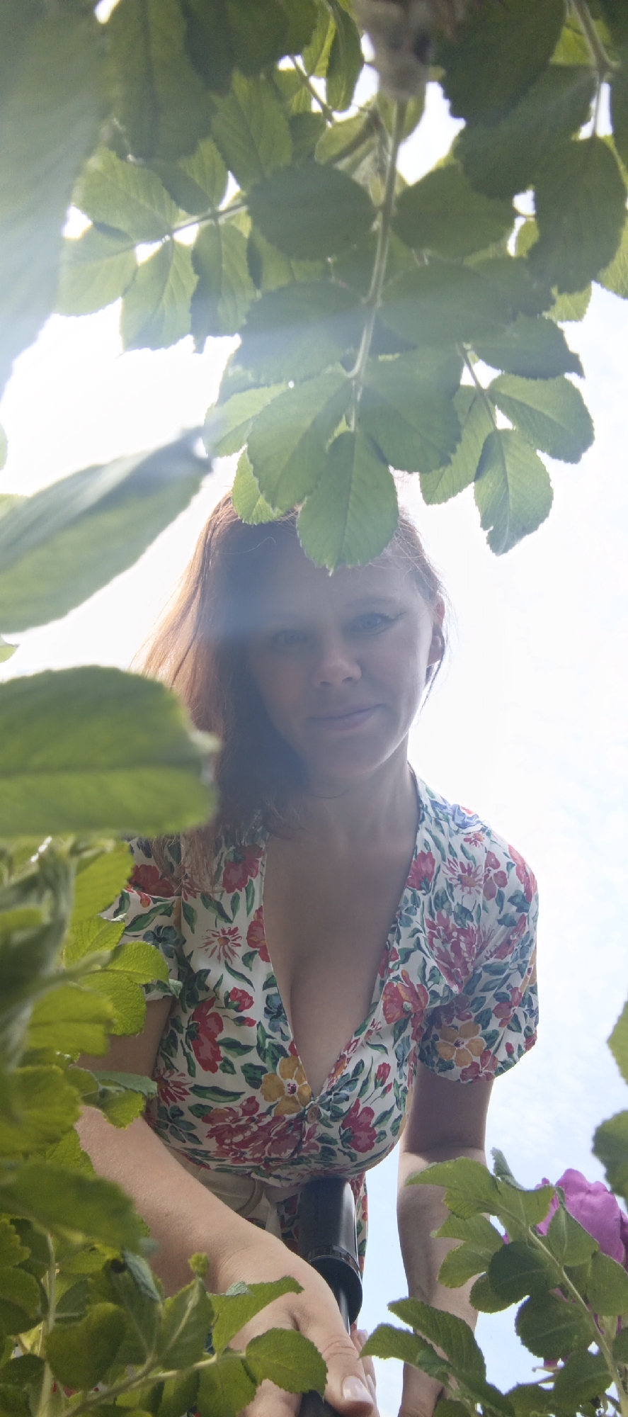 A woman with reddish-brown hair and light skin is looking up at the camera, partially obscured by lush green leaves and branches. She is wearing a floral print dress with a low neckline featuring pink, red, and yellow flowers. Her expression is a soft smile, and her gaze is directed upwards. Her hands are visible, one of which is resting on a dark object, possibly a camera or a support. In the background, the sky is bright and appears to be daytime. The overall impression is that of a candid shot taken outdoors, likely in a garden or natural setting. The location is specified as Chaikovsky, Russia, though no specific landmarks or urban elements are visible to confirm this.