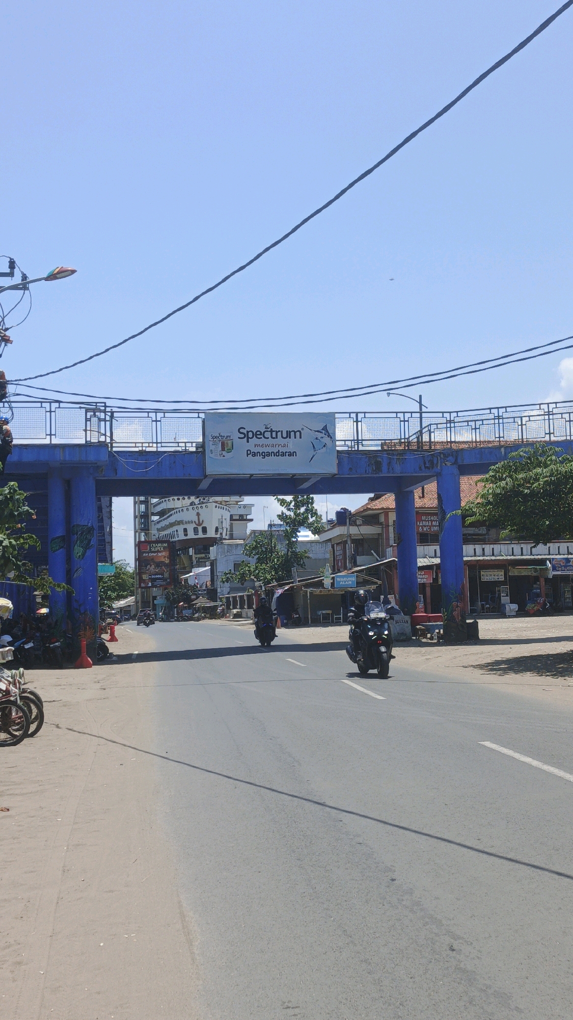 This is a full-frame, eye-level, vertical shot of a street scene in Desa Pagandaran, Indonesia. A blue pedestrian bridge spans the road, adorned with a "Spectrum" advertisement featuring a marlin. Underneath the bridge, a large white building resembling a ship is visible, with other commercial structures lining the street. Two motorcycles are in motion on the asphalt road, one approaching the camera and another further down the road. The sky is clear and bright blue, with power lines crisscrossing the upper portion of the image. The overall scene depicts a sunny day in a coastal urban area.
