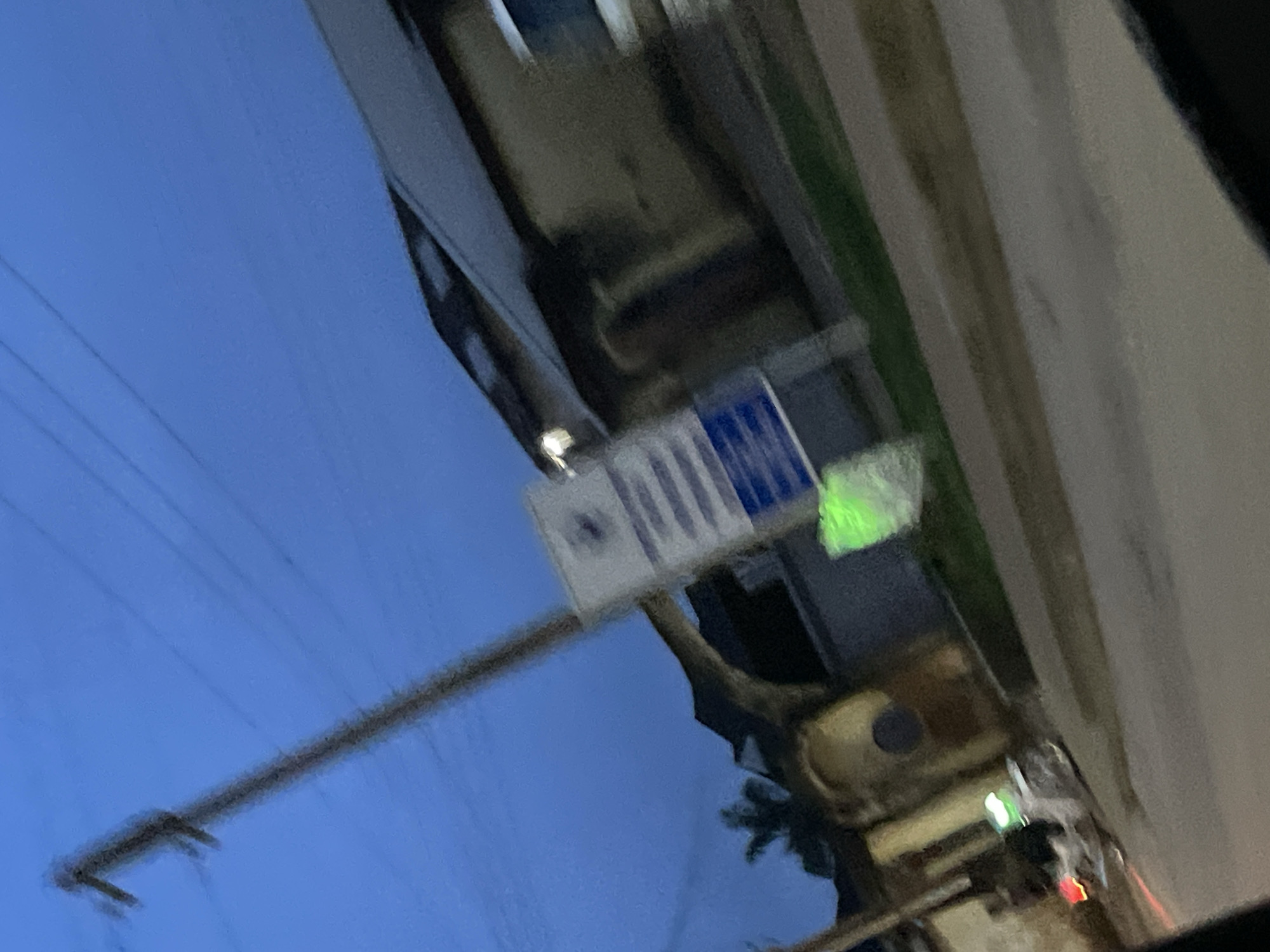 The image depicts an outdoor scene at dusk in Jos, Nigeria. A power pole with overhead wires is prominent on the left. A signpost with blue and white lettering is visible in the mid-ground. To the right, a building with a sloped roof and tan walls is situated. A motorcycle with green and red lights is parked on the right side of the street in the foreground. The street appears to be paved. The sky is a deep blue, indicating twilight.