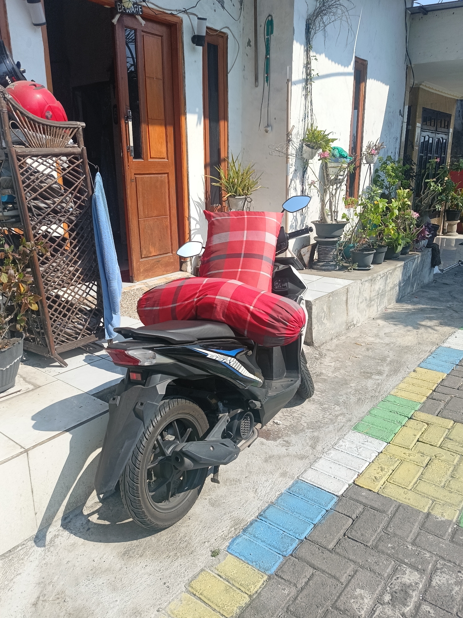 In Gedangan, Indonesia, a red and white checkered bolster pillow and a smaller, similar-patterned pillow are placed on the seat of a black scooter parked on a tiled walkway. The scooter is positioned in front of a house with a wooden door and white walls, adorned with potted plants. To the left of the house, a wooden rack holds a red helmet and a blue towel. The sun casts shadows across the scene.