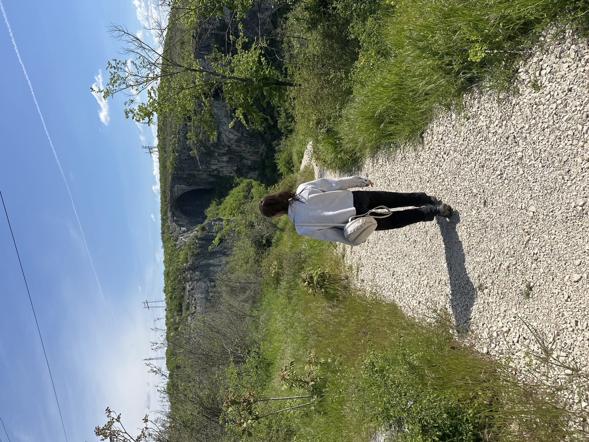 A full-length shot from behind shows a woman walking on a gravel path in Karlukovo, Bulgaria. The woman wears a grey sweatshirt, black pants, and black shoes. She has long brown hair and carries a white purse over her shoulder. She appears to be walking towards a dark tunnel-like structure built into the side of a rocky cliff. The environment includes lush green vegetation and a bright blue sky with wisps of white clouds. Power lines are visible in the background. The path is light-colored gravel. The day appears bright and sunny.