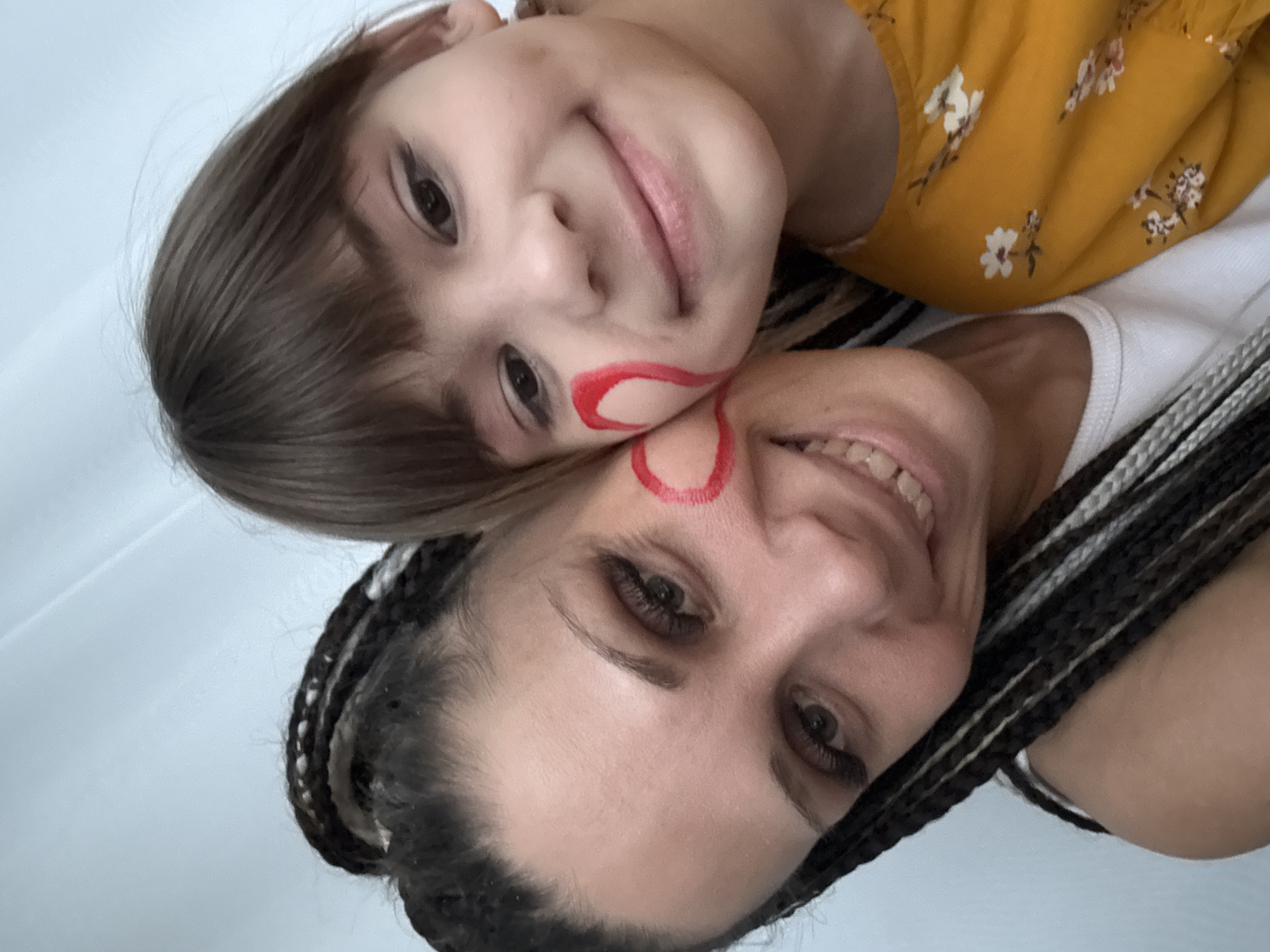 The image is a close-up, overhead selfie featuring an adult woman and a young child, likely a mother and daughter, captured in an intimate and playful moment. Both subjects are fair-skinned and looking up at the camera, smiling.
The child, positioned towards the top of the frame, has straight, light brown hair and dark eyes, with a gentle smile. She is wearing a yellow top with a white floral print. A red heart outline is playfully drawn on her left cheek, extending slightly onto the woman's face.
Below the child is the adult woman, who has dark braided hair that includes lighter streaks. She displays a broad, genuine smile, revealing her teeth, and has dark eyes. A matching red heart outline is drawn on her right cheek, partially overlapping the child's. She is wearing a white tank top visible underneath her arm.
The background is a plain, light-colored surface, suggesting an indoor setting. The lighting is soft and even, indicating either diffused natural light or general indoor illumination. The overall emotion conveyed is one of happiness, affection, and connection between the two individuals, highlighted by their smiles and the shared face paint. The scene evokes a playful family interaction. No text is visible within the image. While no specific location cues are present, the context indicates Rusanovo, Russia.