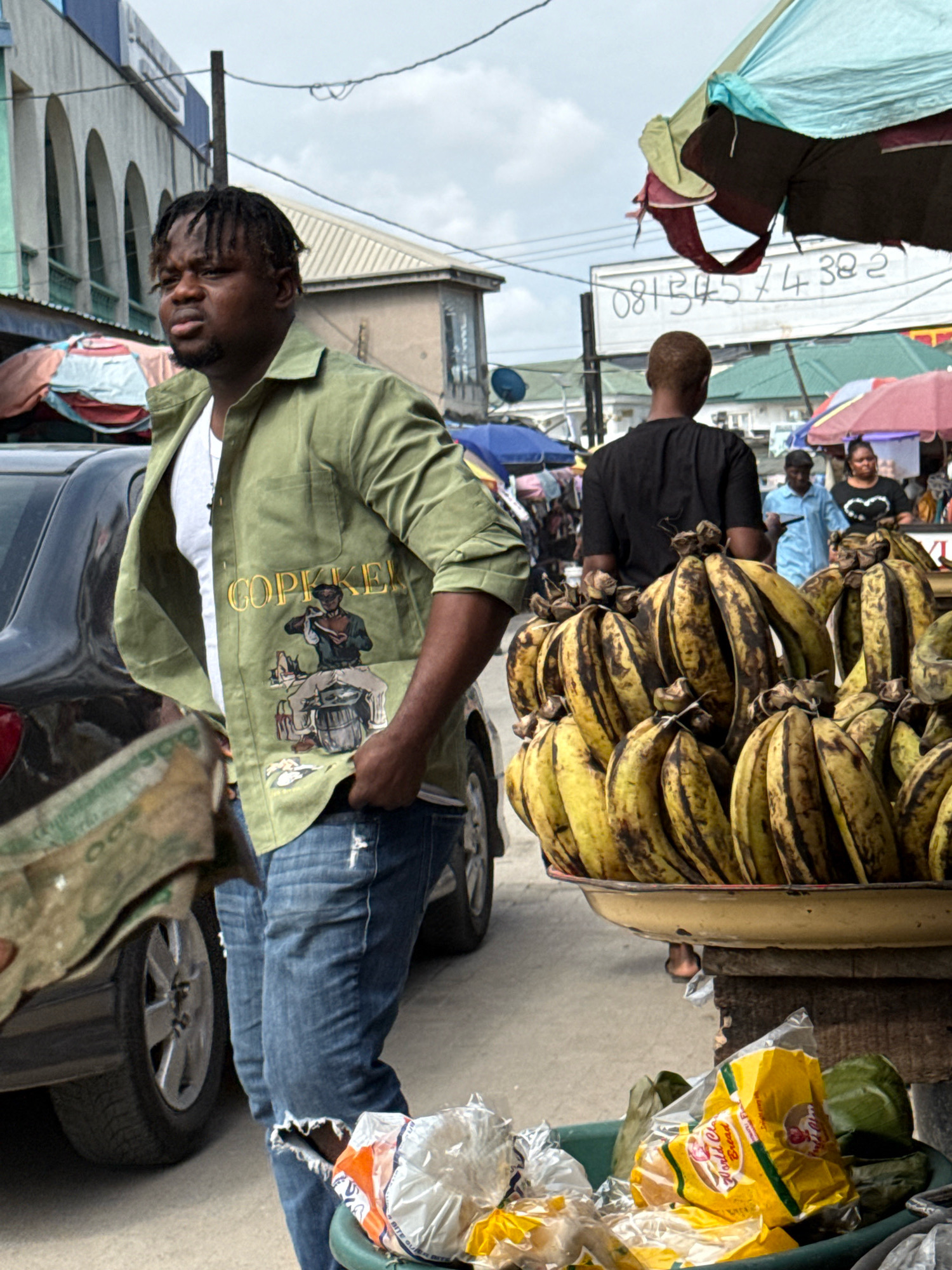 This is a forensic, matter-of-fact description of a street scene in Lekki, Nigeria.
A young man with dreadlocks and a beard, wearing a green button-up shirt with an image and yellow lettering that reads "COPKKEL" and ripped blue jeans, stands on a street. He is holding a wad of Nigerian currency in his left hand. His shirt has an illustration of a person sitting on a barrel. To his right, a large tray is overflowing with ripe plantains, some of which have brown spots. In the foreground, a green plastic basin holds various items wrapped in plastic, including a yellow bag with red lettering that says "World Famous Bread." Behind the man, a dark-colored car is parked. In the background, other market stalls are visible with colorful umbrellas, and several people are walking. A sign with a phone number, "08134574382," is mounted on a pole. The sky is overcast.