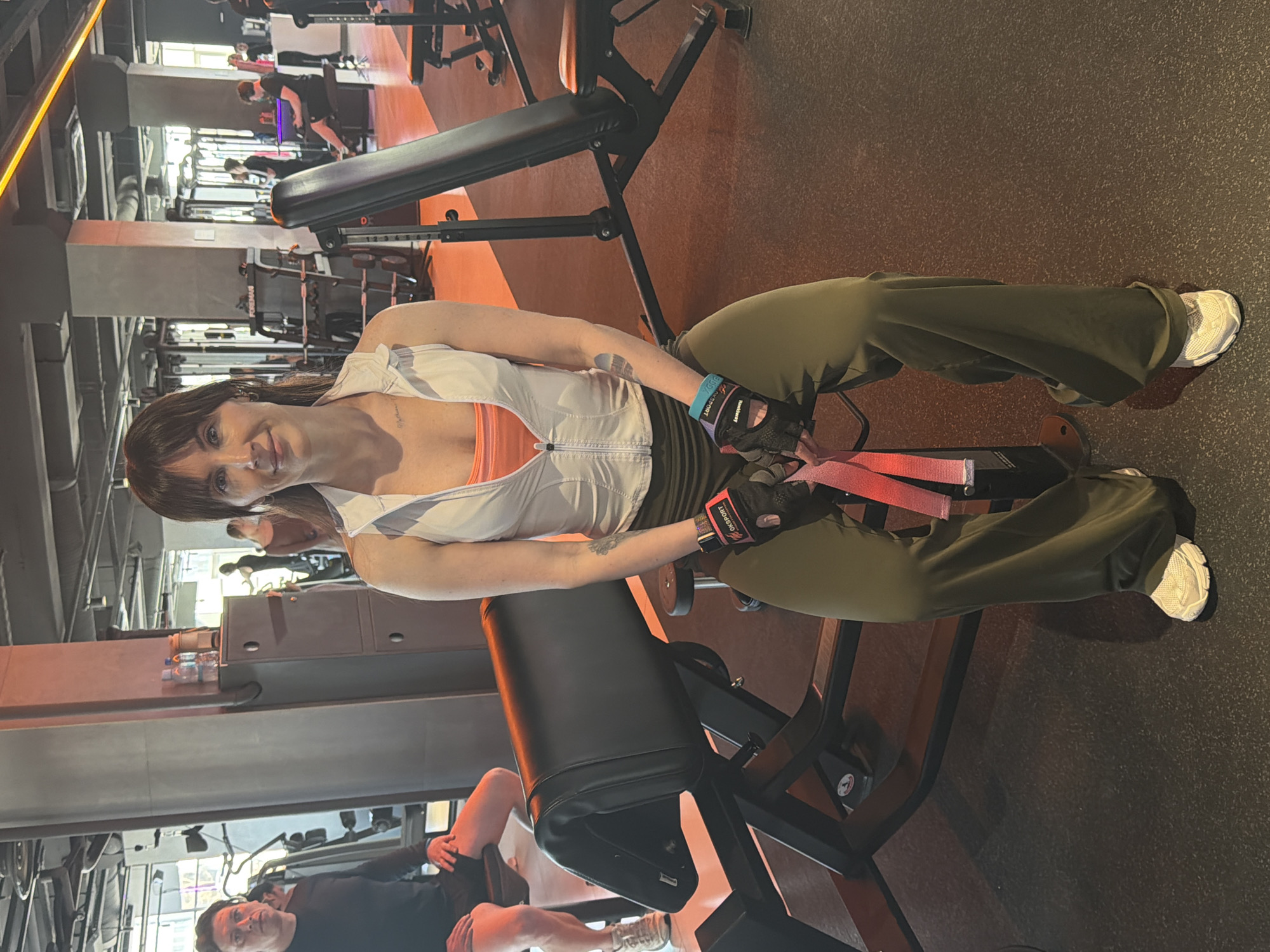 A woman is seated on a gym bench in what appears to be a fitness center in Perm, Russia. She wears a white athletic top, an orange undershirt, olive-green trousers, and white sneakers. She is holding a red resistance band. The fitness center has various exercise machines and other people working out.