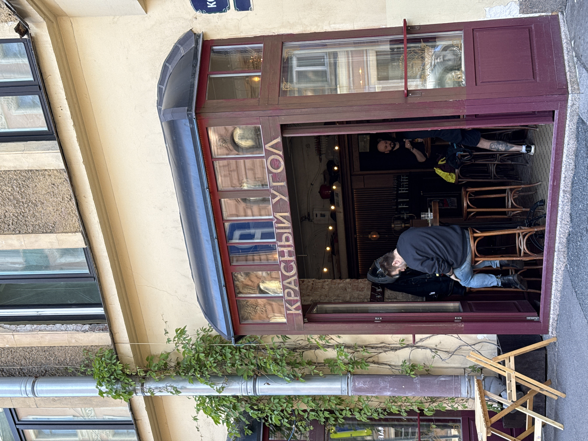 A street-level view captures the entrance to a bar named "КРАСНЫЙ УГОЛ" (Krasny Ugol) in Saint Petersburg, Russia. The building facade is a light yellow with a burgundy-colored entryway featuring glass panels and a black awning. Green ivy climbs a silver downspout to the left of the entrance. Inside the establishment, a man with dark hair sits on a high wooden stool facing away from the camera, wearing a dark sweatshirt and blue jeans. To his right, another man stands by a counter, holding a bottle. A small wooden table and chairs are positioned outside the entrance on the left. The street pavement is visible in the foreground.