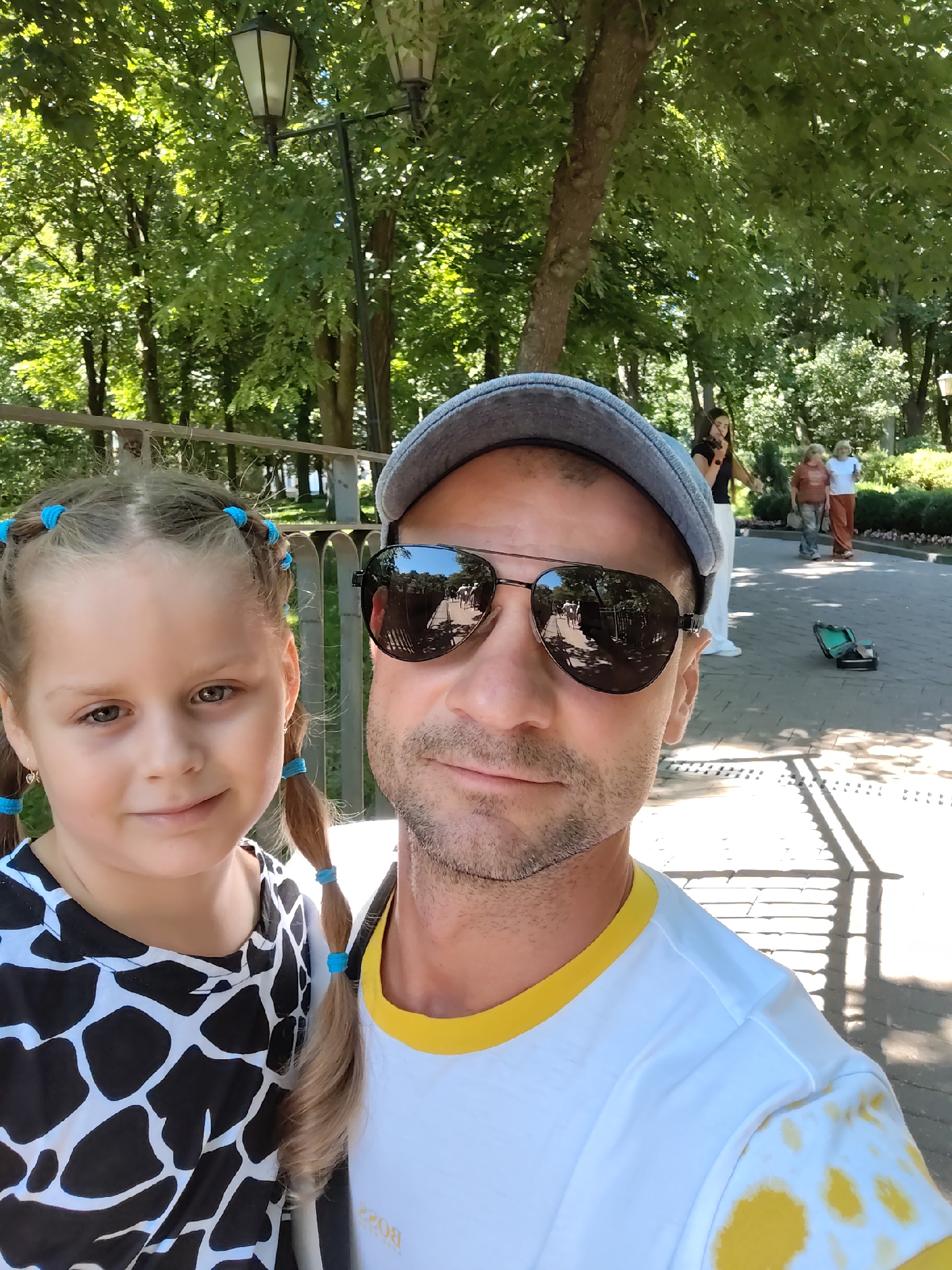 In a park in the city of Homyel, Belarus, a man and a young girl pose for a selfie. The girl, on the left, has her hair in pigtails tied with blue bands and wears a black and white patterned shirt. The man, on the right, wears a grey baseball cap and sunglasses, and a white t-shirt with a yellow collar. The background shows trees, a lamppost, and a pathway where other people are visible, including a woman playing the violin.
