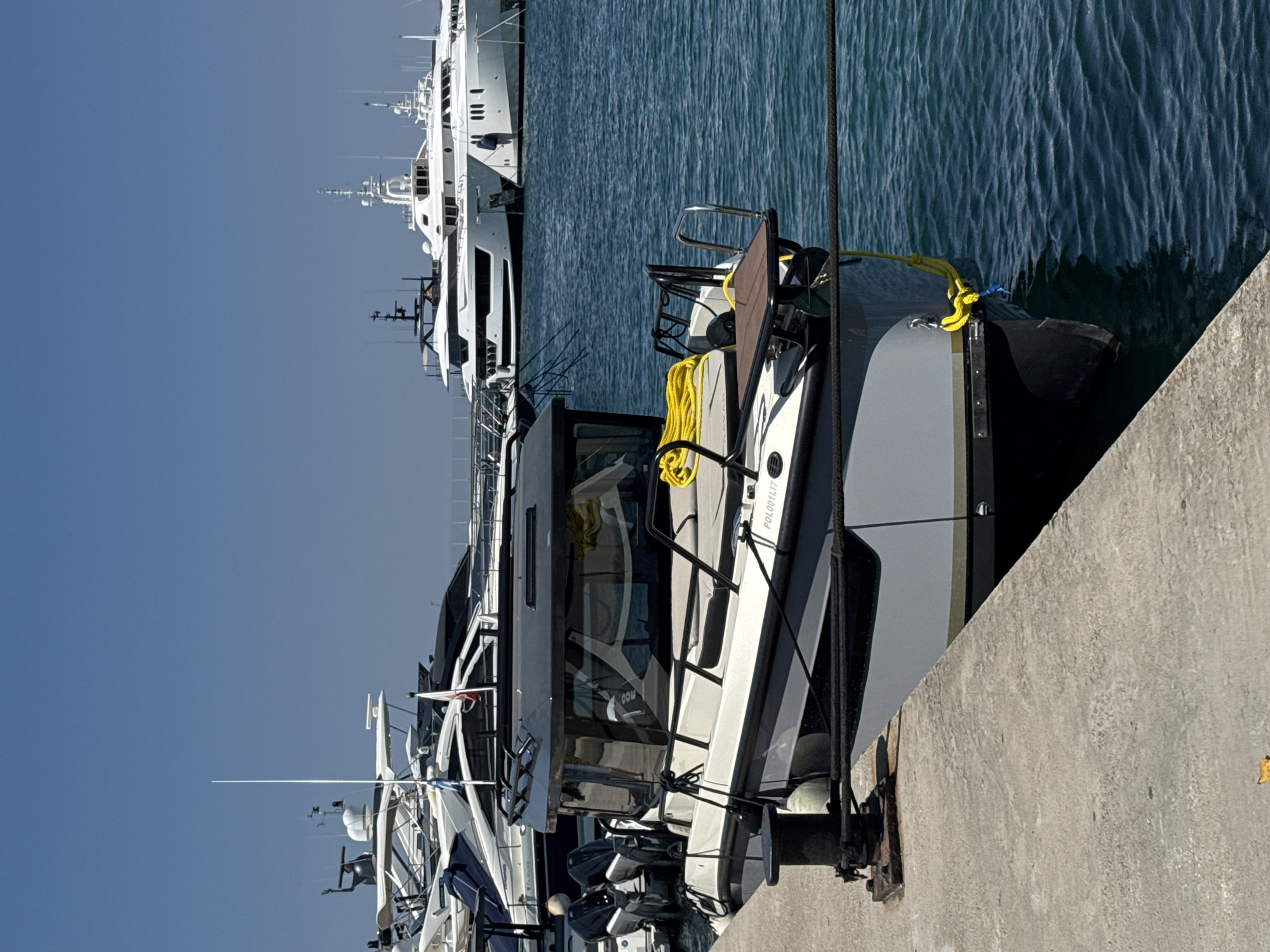 In the city of Moschato, Greece, a medium-sized gray and white motorboat is docked at a concrete pier. The boat has a covered cabin with large windows and twin outboard motors at the stern. A coiled bright yellow rope is visible on the deck, and the boat is secured with dark ropes to a black bollard. In the background, several larger white yachts are moored in the blue water, and a clear blue sky is overhead.
