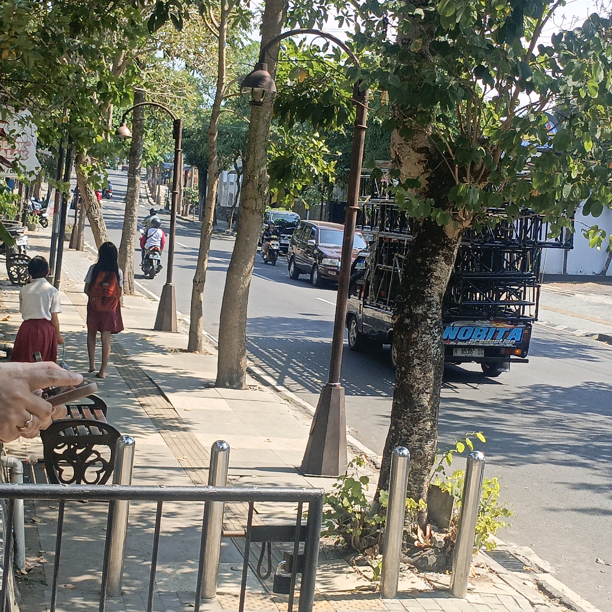 The image captures a street scene in Salatiga, Indonesia, under a bright, sunny sky, casting distinct shadows. On the sidewalk, two young students in uniform, a girl in a white shirt and red skirt, and another girl in a white shirt with an orange backpack and a maroon skirt, are walking away from the camera. One student is holding a mobile phone. Adjacent to the sidewalk, a black metal fence encloses a seating area with a dark metal bench featuring an intricate design.
The street itself is paved and has a gentle incline. Several vehicles are present: a maroon minivan, a motorcycle with a rider wearing a white shirt and a red jacket, and a flatbed truck with a large, dark metal frame structure loaded onto it. The truck bears the word "NOBITA" in blue lettering and license plate "H 8538 IC". Trees line both sides of the street, their branches casting dappled shadows on the pavement and sidewalk. A vintage-style street lamp with a bell-shaped fixture stands near the sidewalk. The environment appears to be a well-trafficked urban or suburban area.