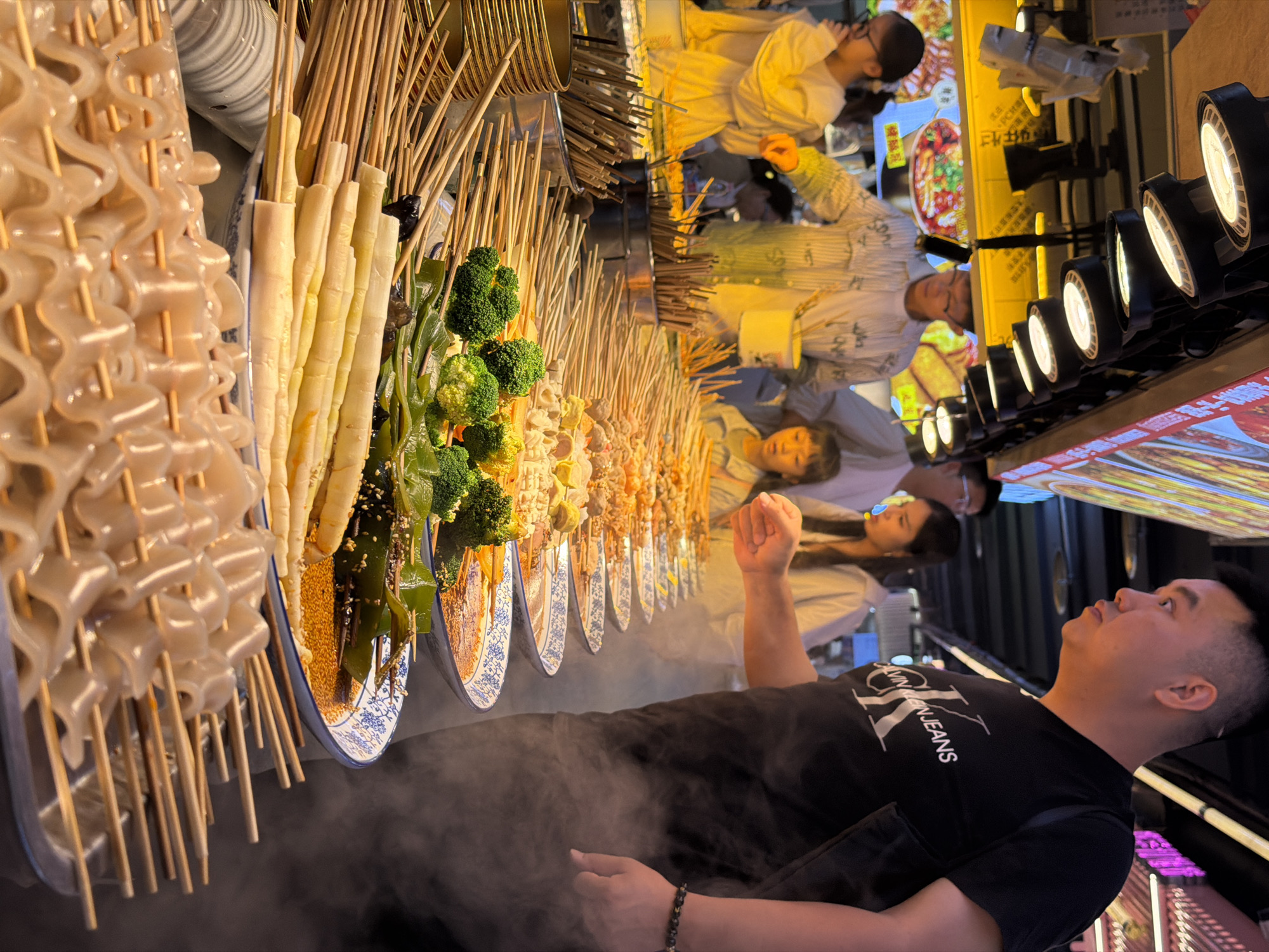 A street food stall in Yuzhong District, China displays rows of skewered food items, including broccoli, bamboo shoots, and a pale, wavy gelatinous food, arranged in bowls of broth. A man in a black t-shirt with a "Calvin Klein Jeans" logo is visible on the right, looking towards the food and a woman with long dark hair in a white shirt on the left. Several other people, including a young girl in a light-colored top, are present in the background. The stall is illuminated by overhead lights, and steam rises from the food.