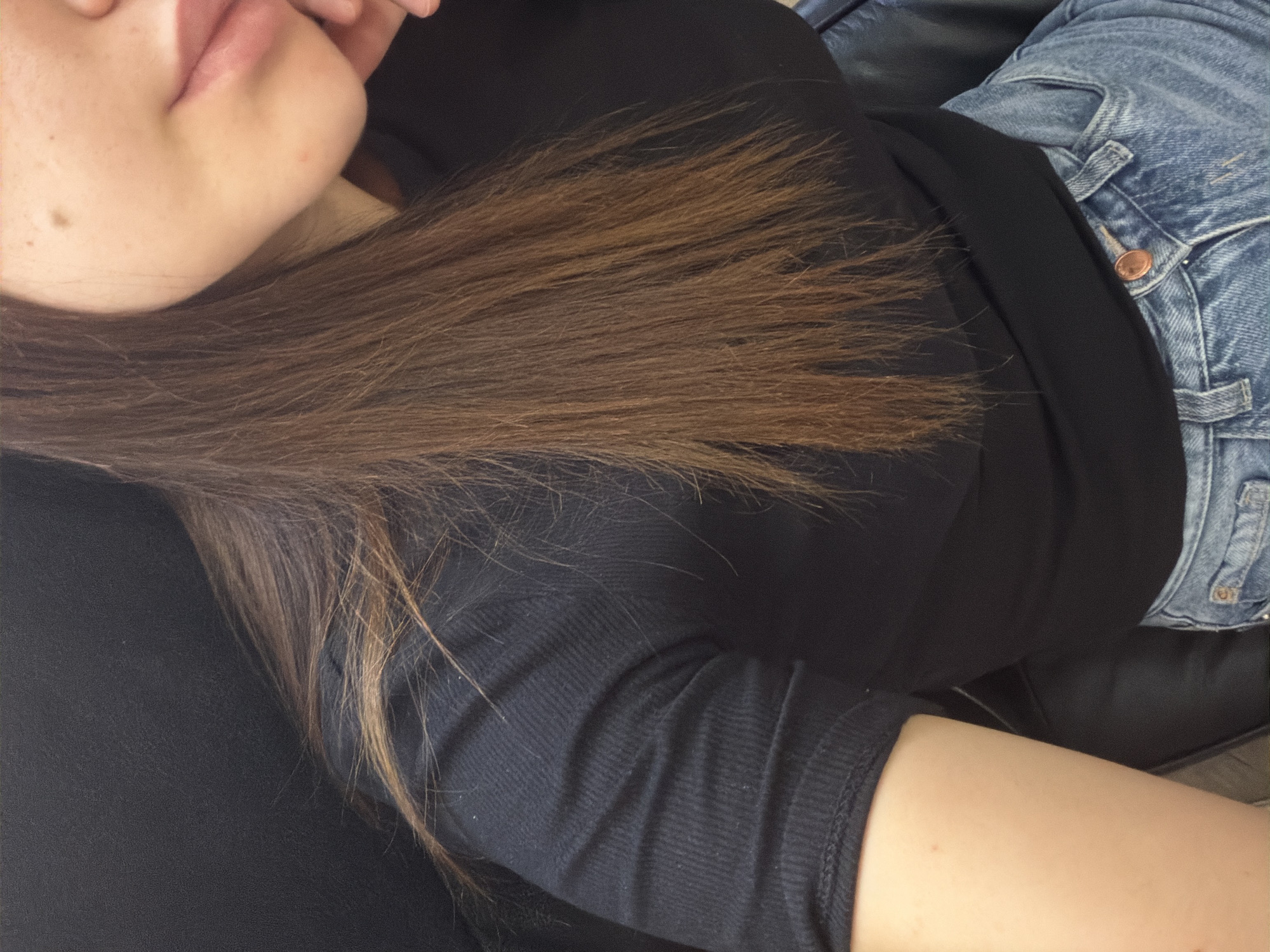 This is a close-up, medium-shot photograph of a person's upper body. The subject is wearing a black t-shirt and blue jeans. Their right arm is visible, leaning on a dark surface, possibly a chair. The person's long, brown hair is parted and falls over their shoulder and chest. The lower part of their face, including their lips and chin, is visible on the right side of the frame. The background is dark and indistinct, with no discernible city or environmental features. The photograph does not provide enough information to determine its location in Chișinău, Moldova, or any specific actions or interactions.