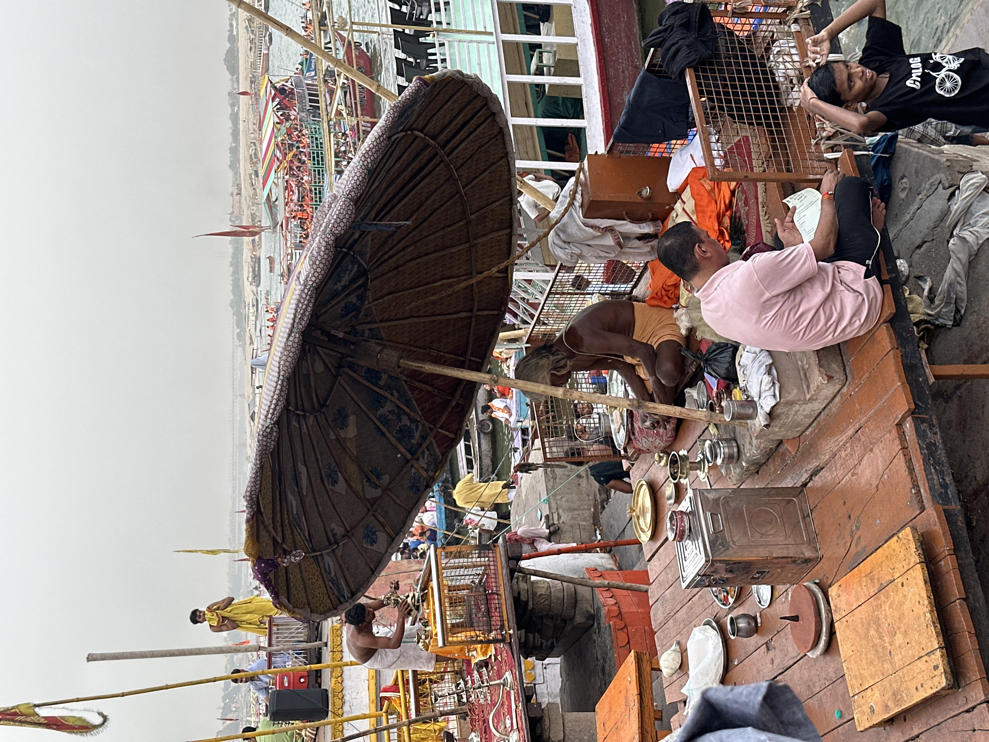 The image depicts a scene on the ghats of Varanasi, India.
Several individuals are present. A man in a pink shirt sits on a wooden platform, looking at papers with a young boy lying next to him. Another man, shirtless and wearing orange attire, is attending to something on the ground. A partially visible individual is wearing a yellow garment. In the background, numerous people gather near boats on the riverbank.
An aged umbrella provides shade, and a variety of objects such as metal containers, plates, and other paraphernalia are scattered on the platform. The setting is riverside with a view of the Ganges River, lined with boats and buildings. The sky is overcast.
