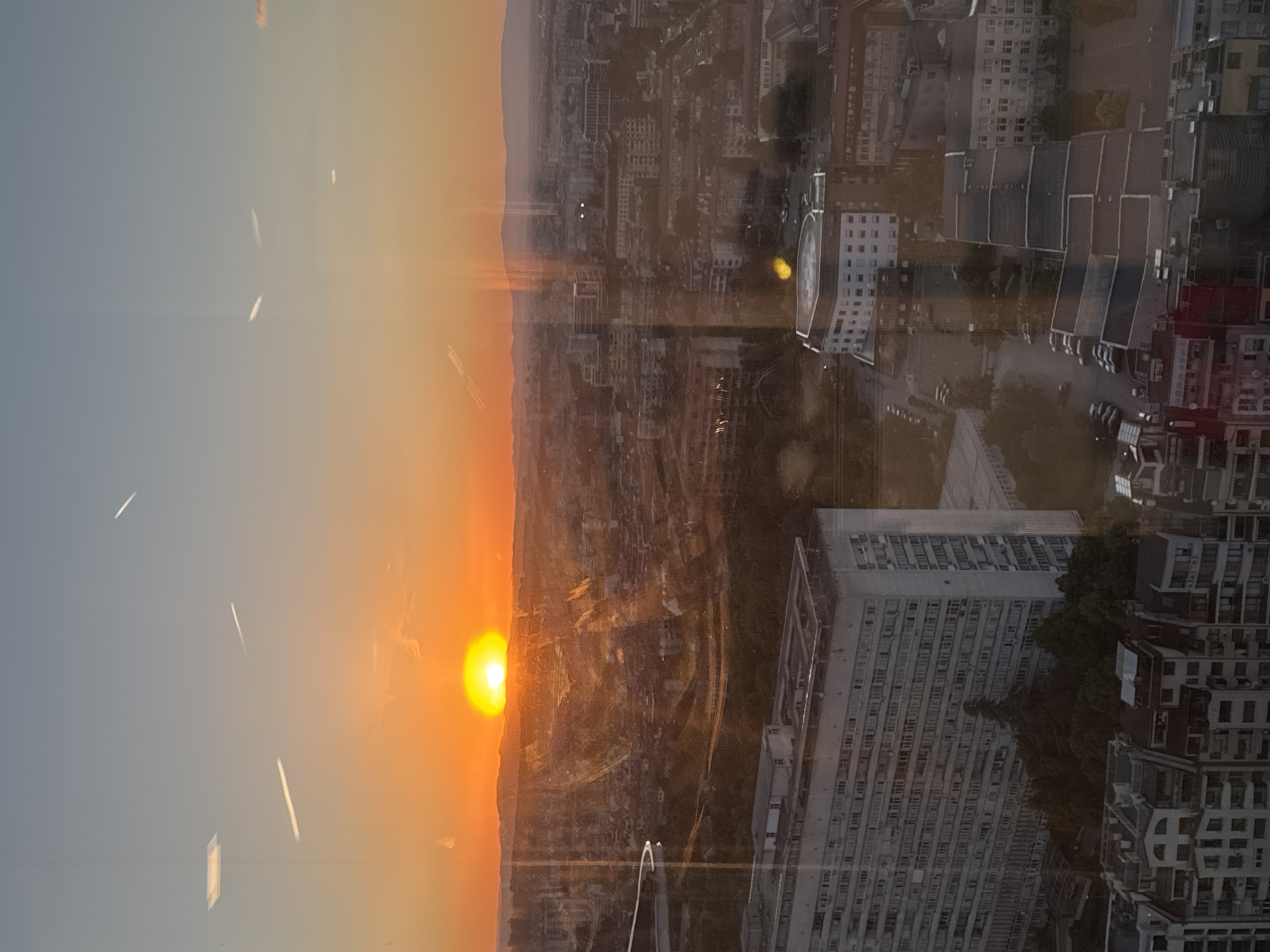 The image presents a rotated view from an elevated position, possibly a high-rise building, overlooking a cityscape during sunset. The sun is situated on the left, emitting an orange glow that permeates the sky and reflects off the urban landscape. The city itself comprises a dense collection of buildings, ranging in height and style, with visible streets and possible traffic activity. The environment appears to be Sofia, Bulgaria, identified by the building's architecture. The image captures a moment in time where natural light interacts with the urban environment, creating a contrast between the vibrancy of the sunset and the architectural forms below. The image seems to have been taken through a window, as streaks are visible.