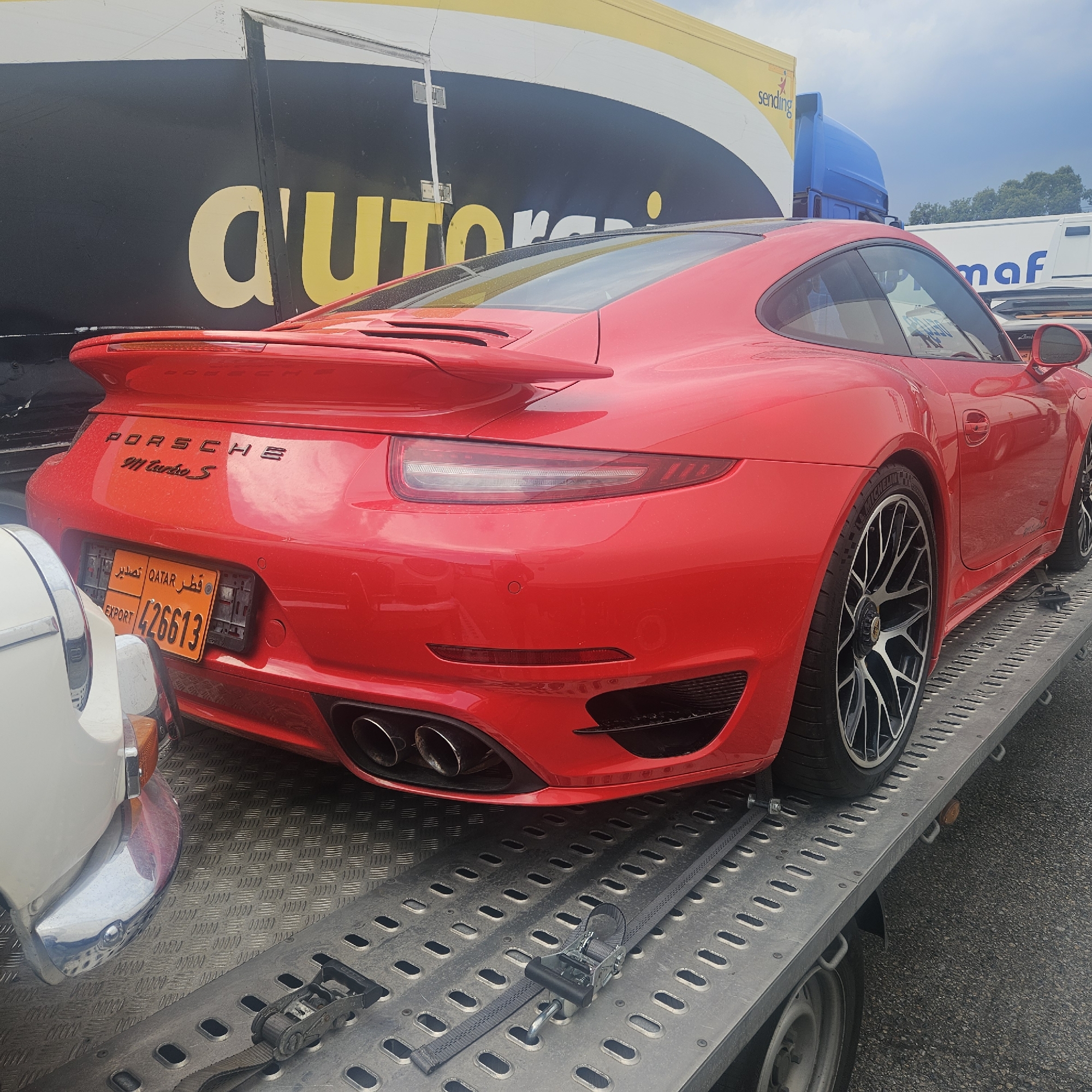 The image shows a red Porsche 911 Turbo S on a trailer, parked on asphalt. The car has a black spoiler and black wheels. A license plate from Qatar is visible on the rear of the car, with the number 426613. To the left of the Porsche, a portion of a white classic car is visible. In the background, there is a large black and yellow truck with the word "AUTOR" visible. Further back, a blue truck and a white truck with the word "maf" are also present. The sky is overcast. The scene is consistent with a vehicle transport location, possibly a parking lot or depot, in the vicinity of La Jonquera, Spain, a common location for international vehicle transport due to its proximity to the French border. No people are visible in the image.